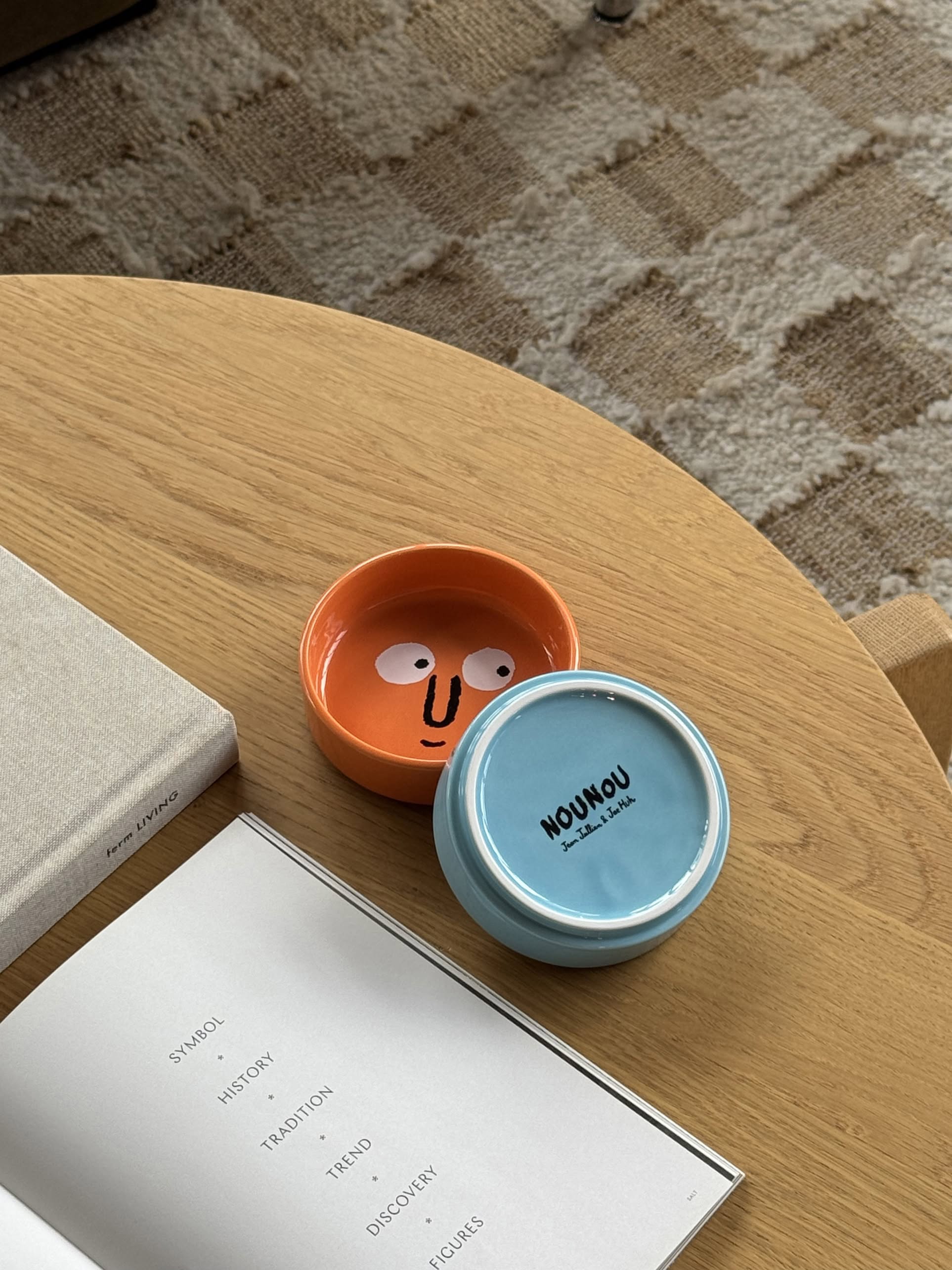A round wooden table holds an open book, a beige hardcover, a NouNou Friends Plate Set in Mint Blue/Coral Orange, and a blue bowl labeled HOUNOU BY MILKLAB. A patterned rug lies below.