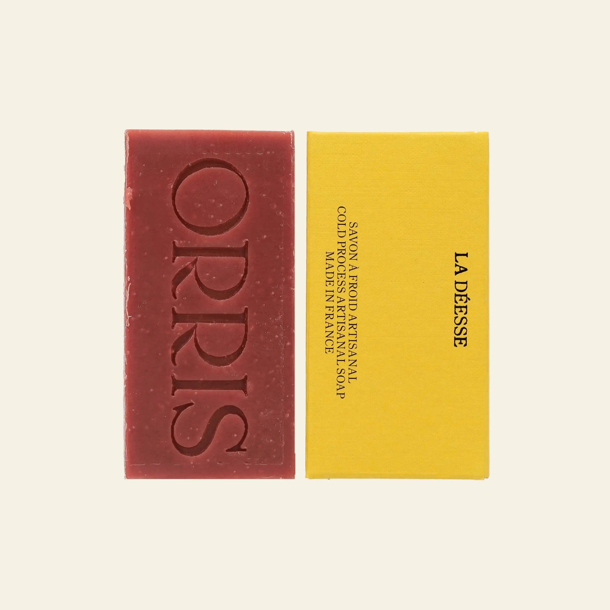 A rectangular red ORRIS La Déesse Botanical Soap bar, with ORRIS embossed on it and containing Ayurvedic Manjistha, sits beside a yellow box labeled La Déesse in black text. The scene is set against an off-white background.