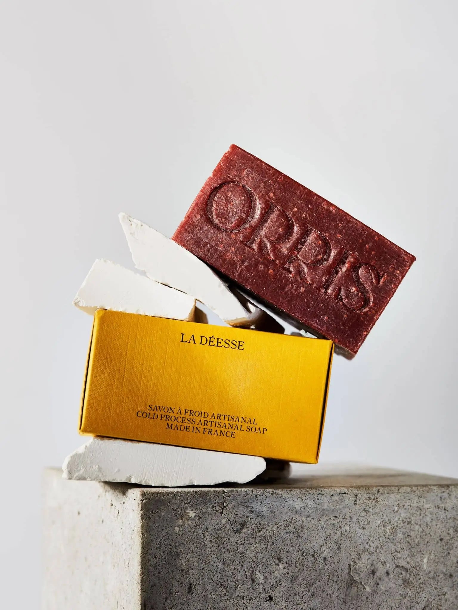 Three white soap pieces and one dark red ORRIS Botanical Soap, embossed with ORRIS, are stacked on a yellow ORRIS La Déesse box, set on concrete with a neutral backdrop—a collection evoking Ayurvedic Manjistha and raw honey.