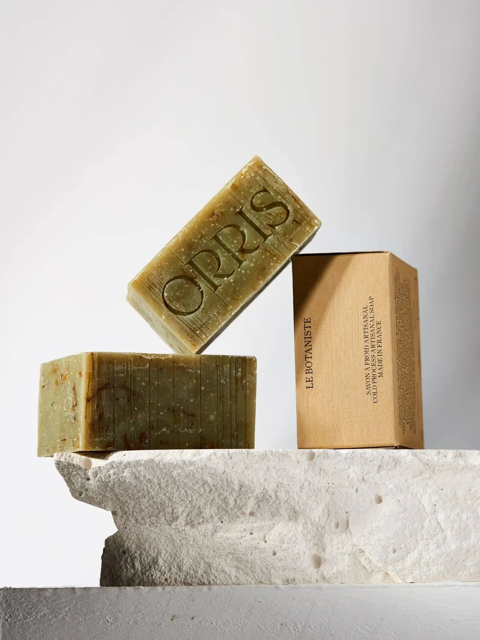 Three ORRIS Le Botaniste handcrafted natural soap bars are displayed on a rough white stone: one lies on its side, one stands upright, and the third is boxed in brown packaging with botanical text. One bar features an ORRIS engraving.