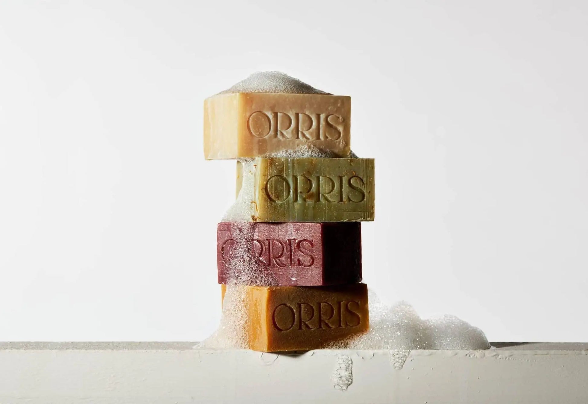 Four stacked bars of ORRIS Le Botaniste botanical soap in assorted colors, topped with white bubbles. One bar displays calendula petals for a natural touch, all set against a plain white background. Brand: ORRIS.