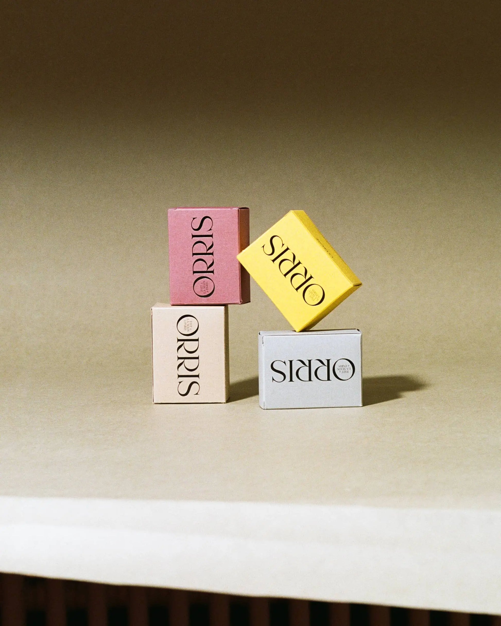 Three small handcrafted ORRIS Le Botaniste soap boxes in pink, yellow, and light gray are stacked in a minimalist setting. The botanical boxes—by ORRIS, including Calendula—are arranged in an elegant, balanced composition.