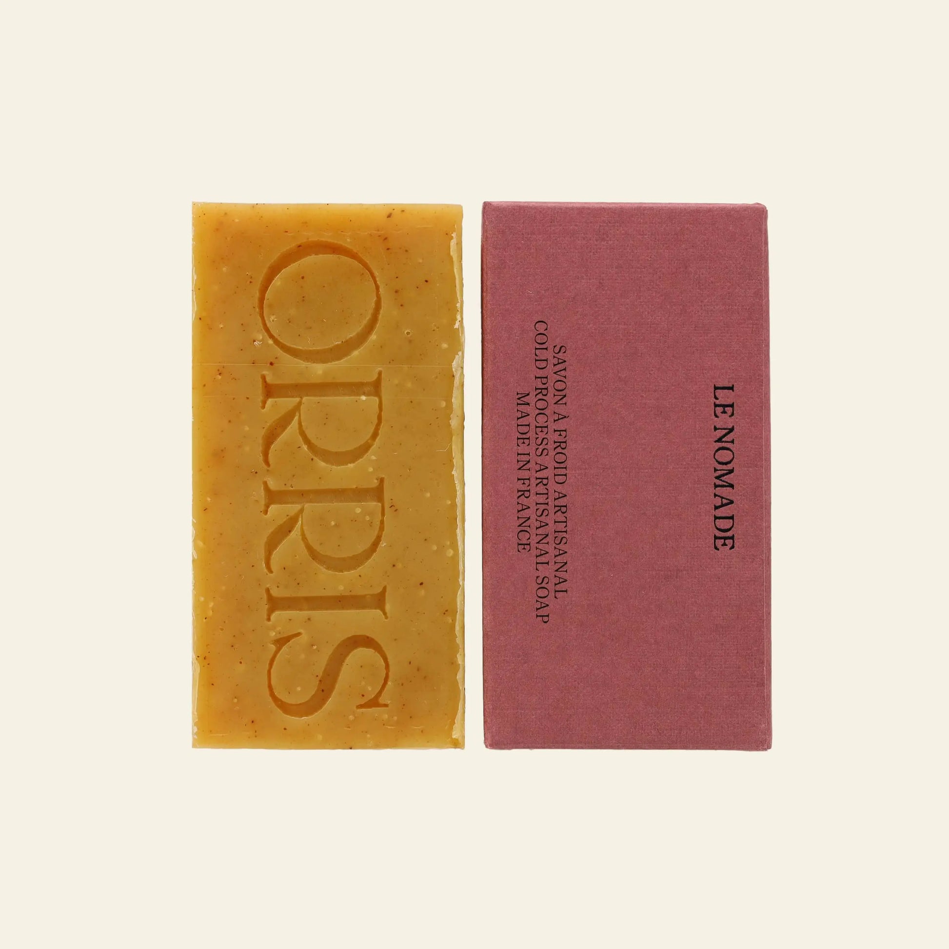 Two rectangular bars of ORRIS Le Nomade botanical soap rest side by side on a light background—one tan with ORRIS embossed and the other in mauve packaging—showcasing the fine French craftsmanship of ORRIS.