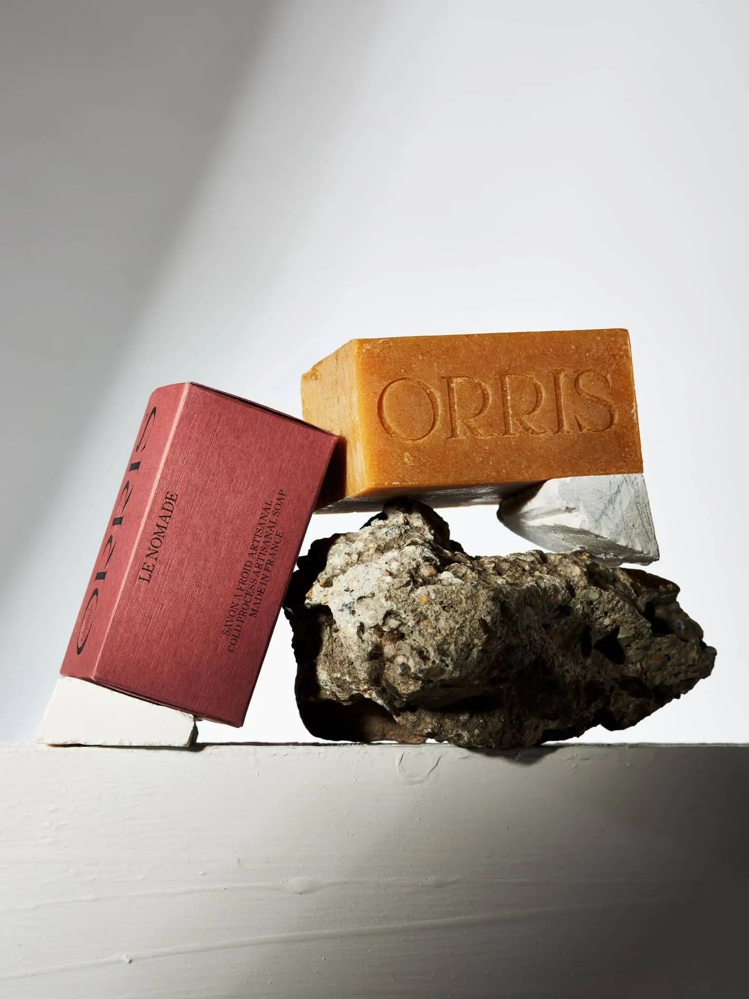 A bar of ORRIS Le Nomade artisanal botanical soap, embossed with ORRIS and paired with a red box, leans against a rough stone on a white surface under dramatic lighting and shadows.