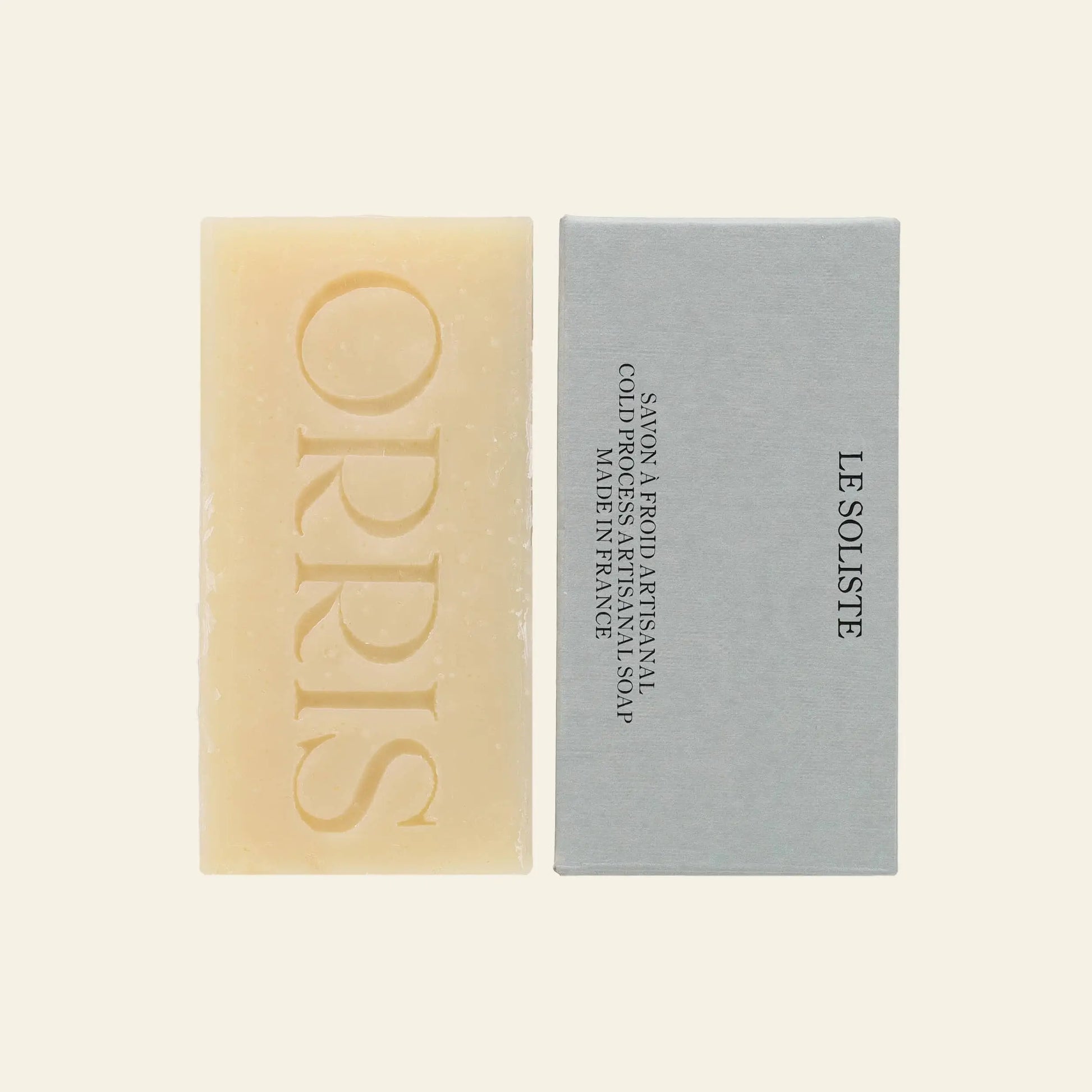 A rectangular beige ORRIS Le Soliste artisanal soap bar, embossed with ORRIS, rests beside its light grey packaging labeled LE SOLISTE. The cruelty-free bar is set against a plain light background.