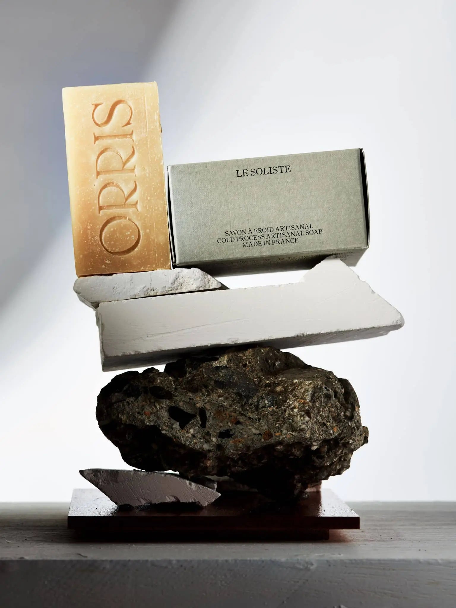 An ORRIS Le Soliste soap bar and its box are artfully balanced on stone and plaster, forming a sculptural display on a flat surface with soft shadows. This cruelty-free ORRIS product elevates the scene.