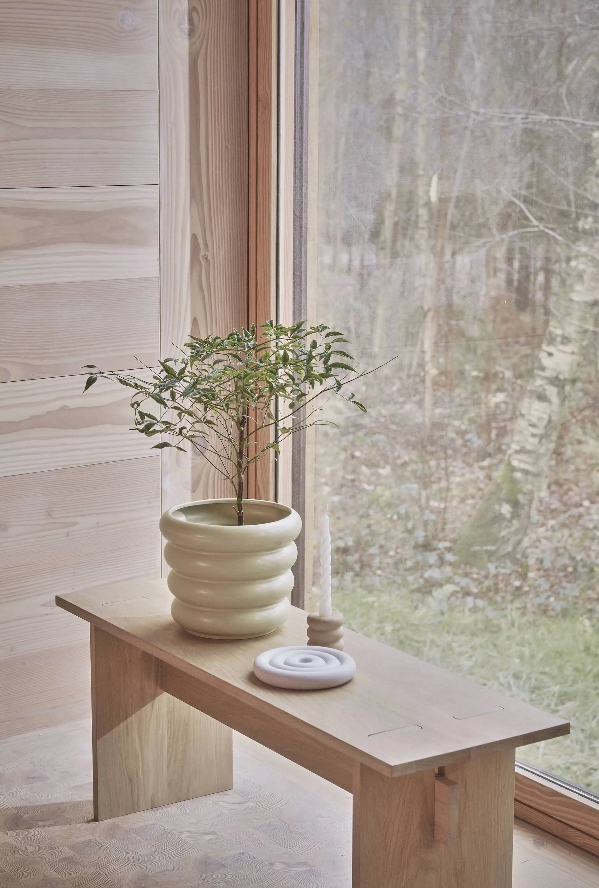 A minimalist wooden bench by the window holds a leafy plant in the OYOY Living Design Awa Pot, Large, alongside a white taper candle holder and a round white tray, creating a botanical scene that echoes the outdoor greenery.