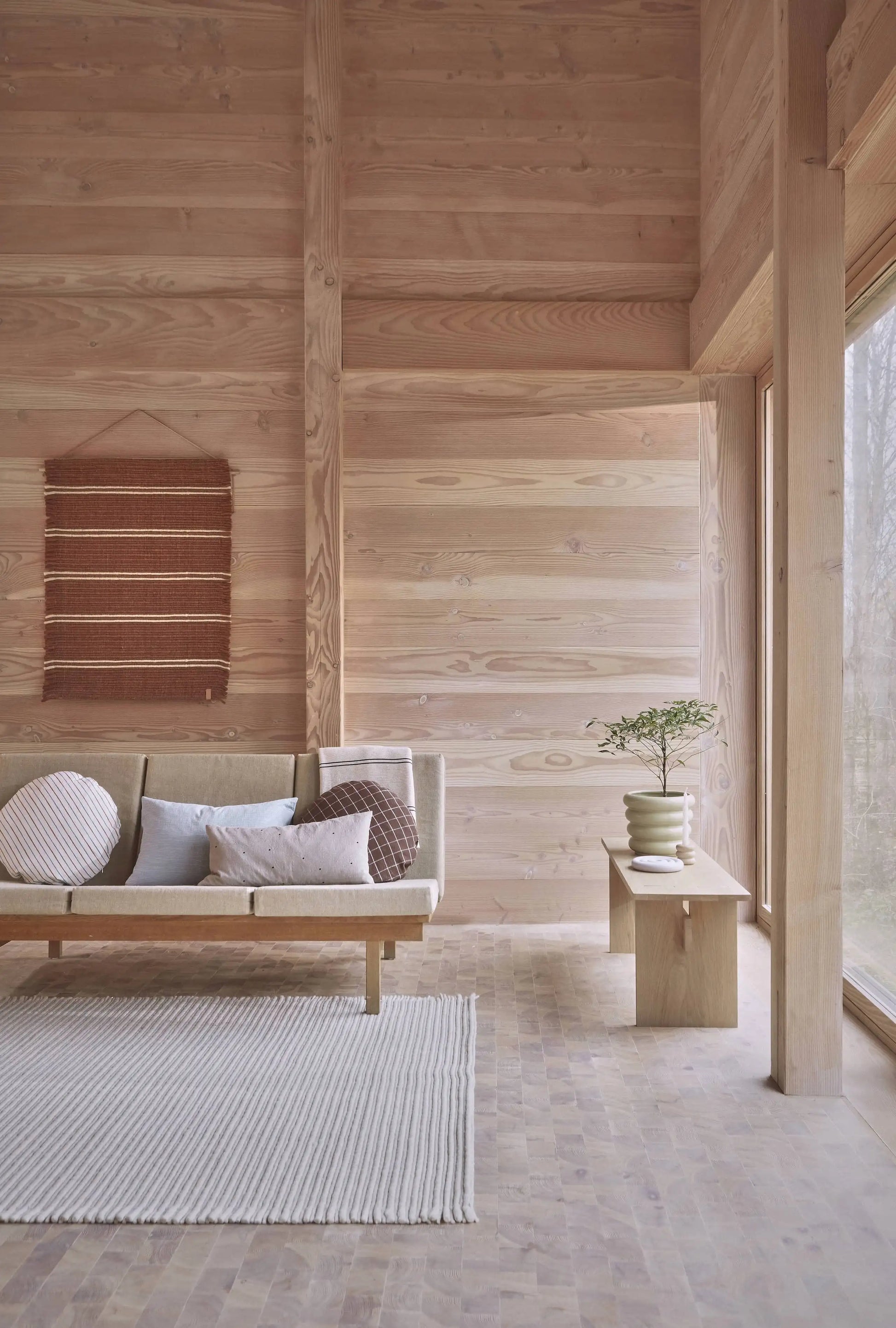 Minimalist living room with light wood walls, a beige sofa, textured white rug, and wooden bench showcasing the OYOY Living Design Awa Pot, Large filled with greenery beside large windows; a woven wall hanging completes the natural look.