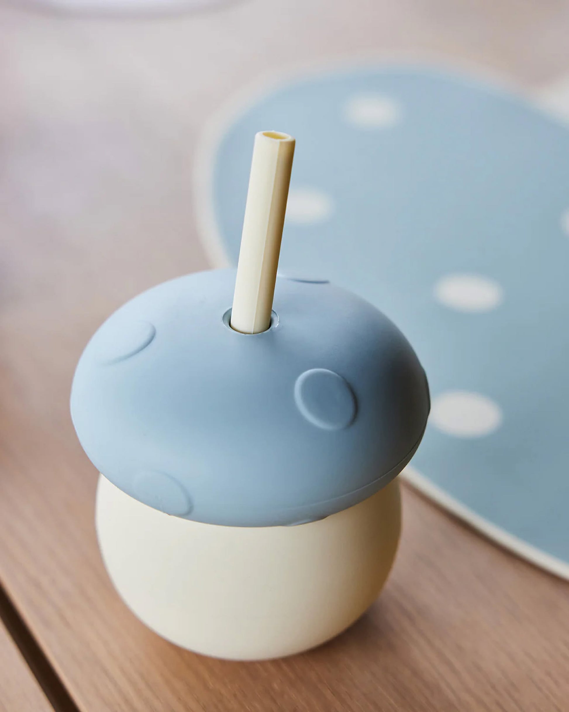 The OYOY Living Design Mushroom Cup, featuring a blue mushroom-shaped lid and straw, rests on a wooden surface with a blurred blue polka-dotted mat in the background.