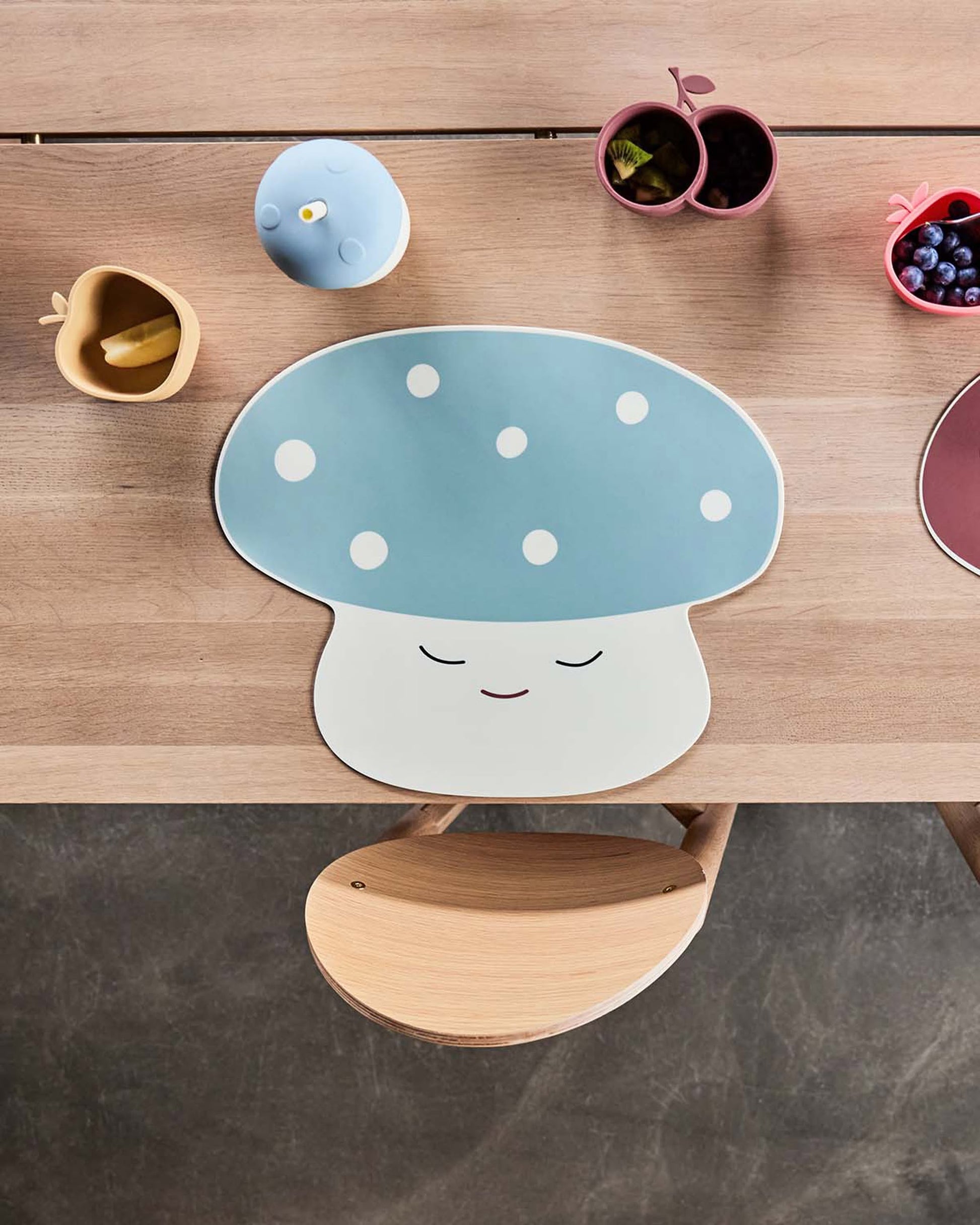 A wooden table set with a blue and white OYOY Living Design Mushroom Cup, small apple- and pear-shaped bowls filled with fruit and snacks, a wooden chair, and a cheerful, child-friendly atmosphere.