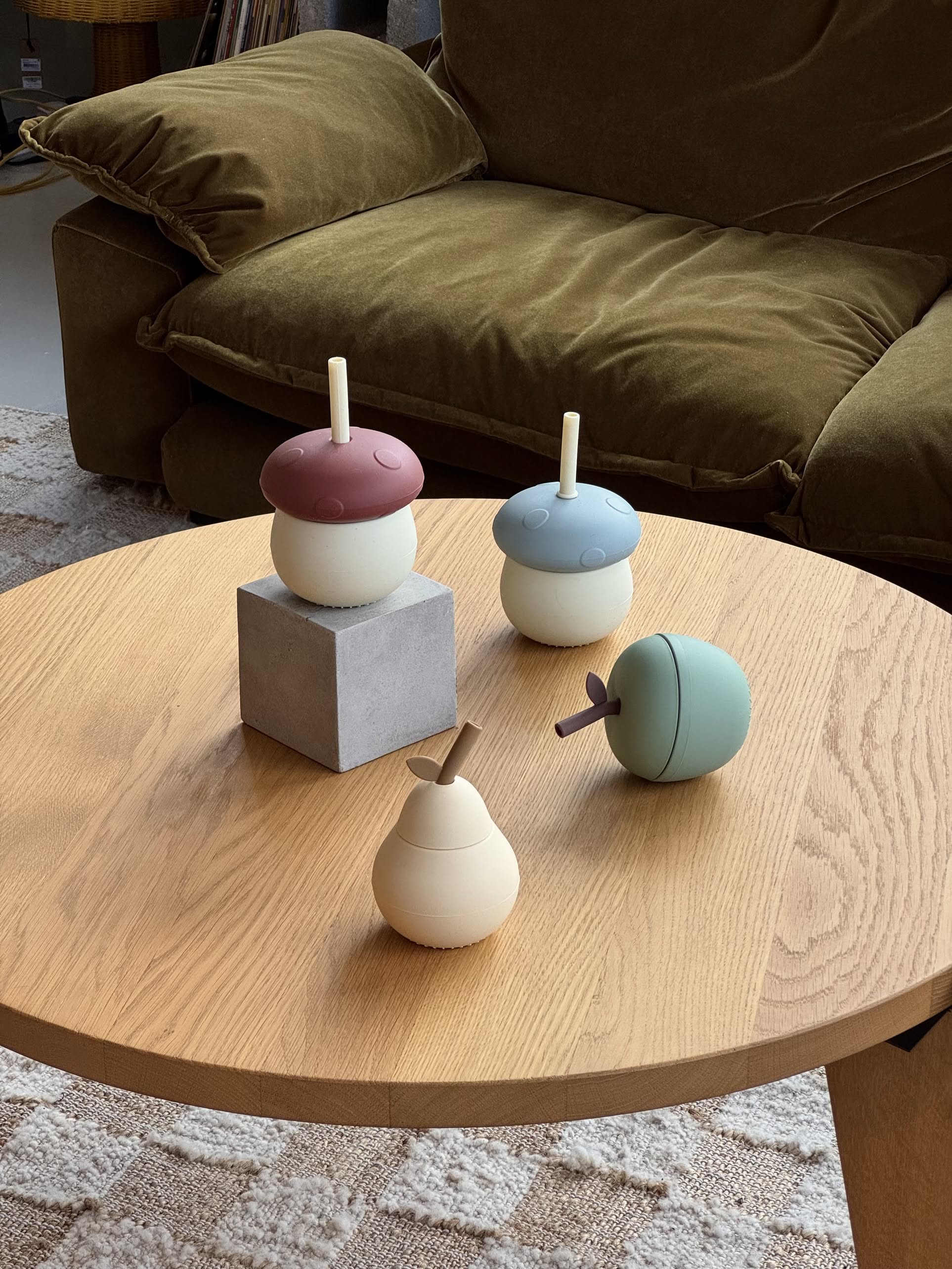 The OYOY Living Design Mushroom Cup, along with three playful silicone cups shaped like fruits and an acorn, are displayed on a round wooden coffee table in front of a green couch; one cup sits atop a grey block on a textured rug.