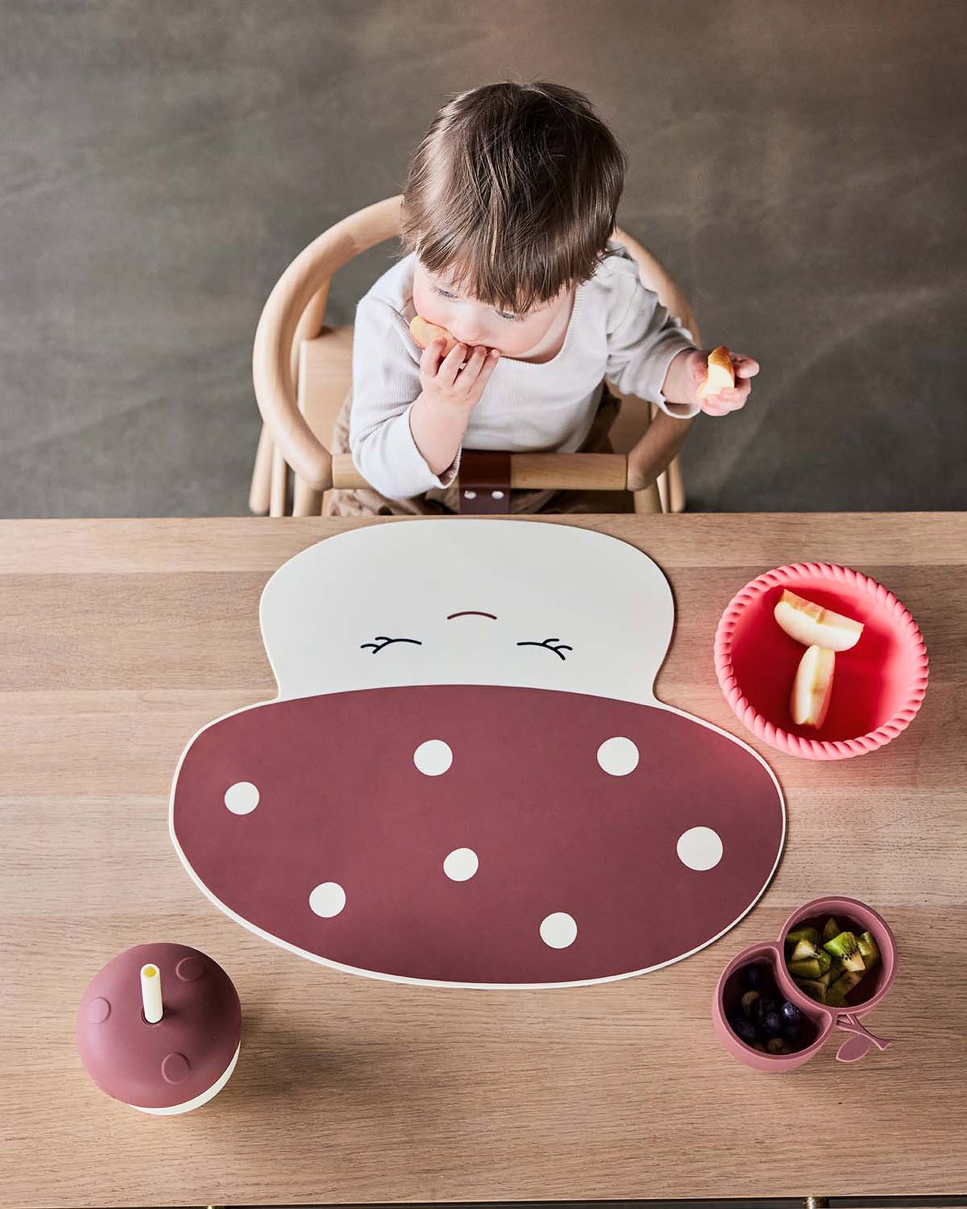 A small child sits at a wooden table eating a snack. In front of them is a cute polka-dot placemat, a red bowl with banana pieces, and the OYOY Living Design Mushroom Cup with a straw beside a small dish of grapes and blueberries.