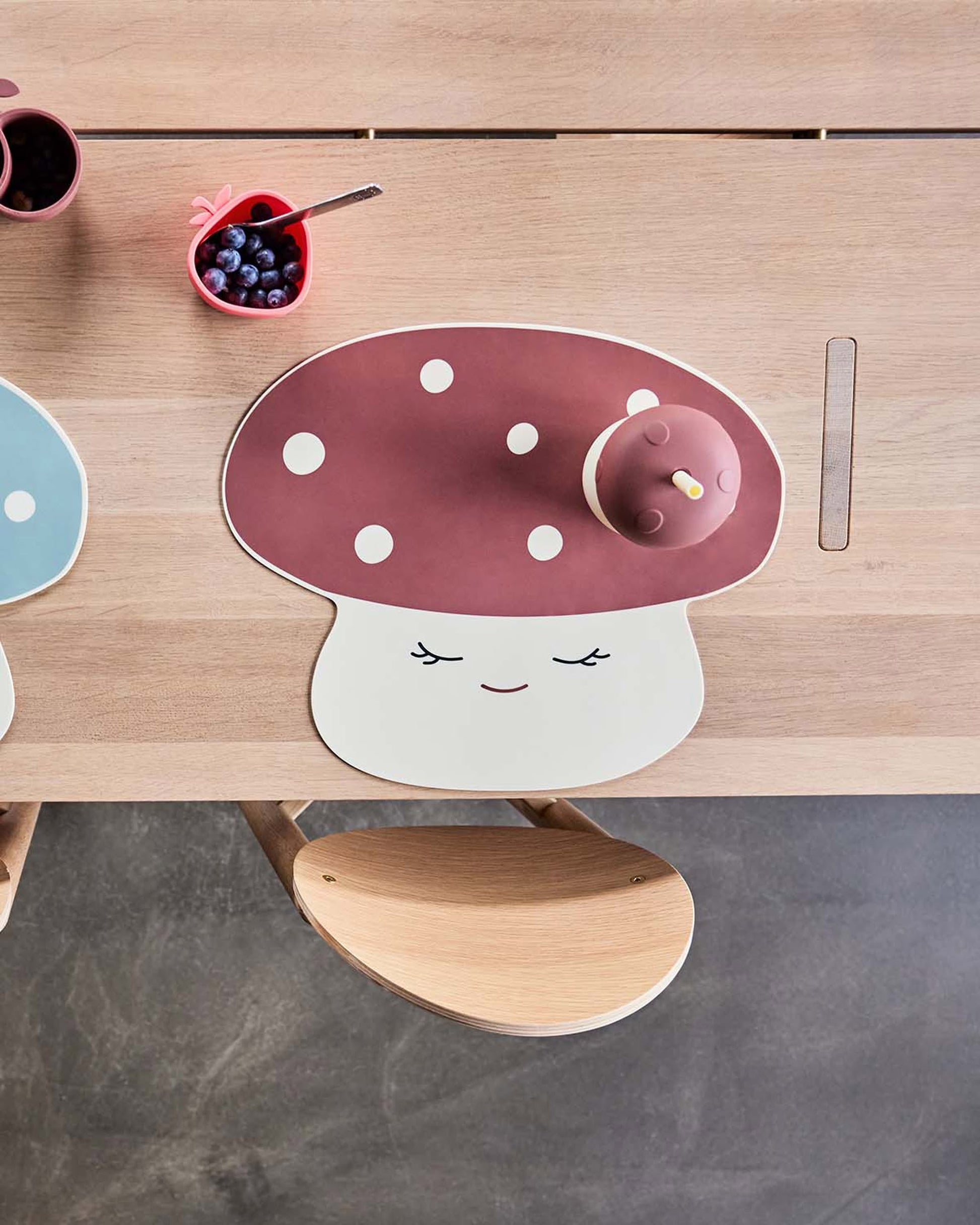 A wooden table set with a cheerful mushroom-shaped placemat, the OYOY Living Design Mushroom Cup with lid, and a pink heart-shaped dish of blueberries. A light wooden chair from OYOY Living Design is partially visible at the table.