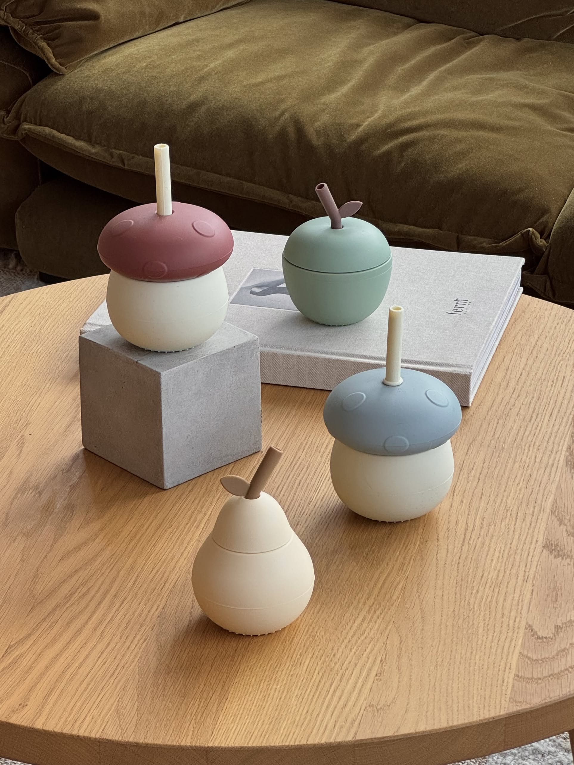 Four playful OYOY Living Design Mushroom Cups with lids and straws sit on a wooden table, one perched on a gray cube, with two books in the background near a brown couch.
