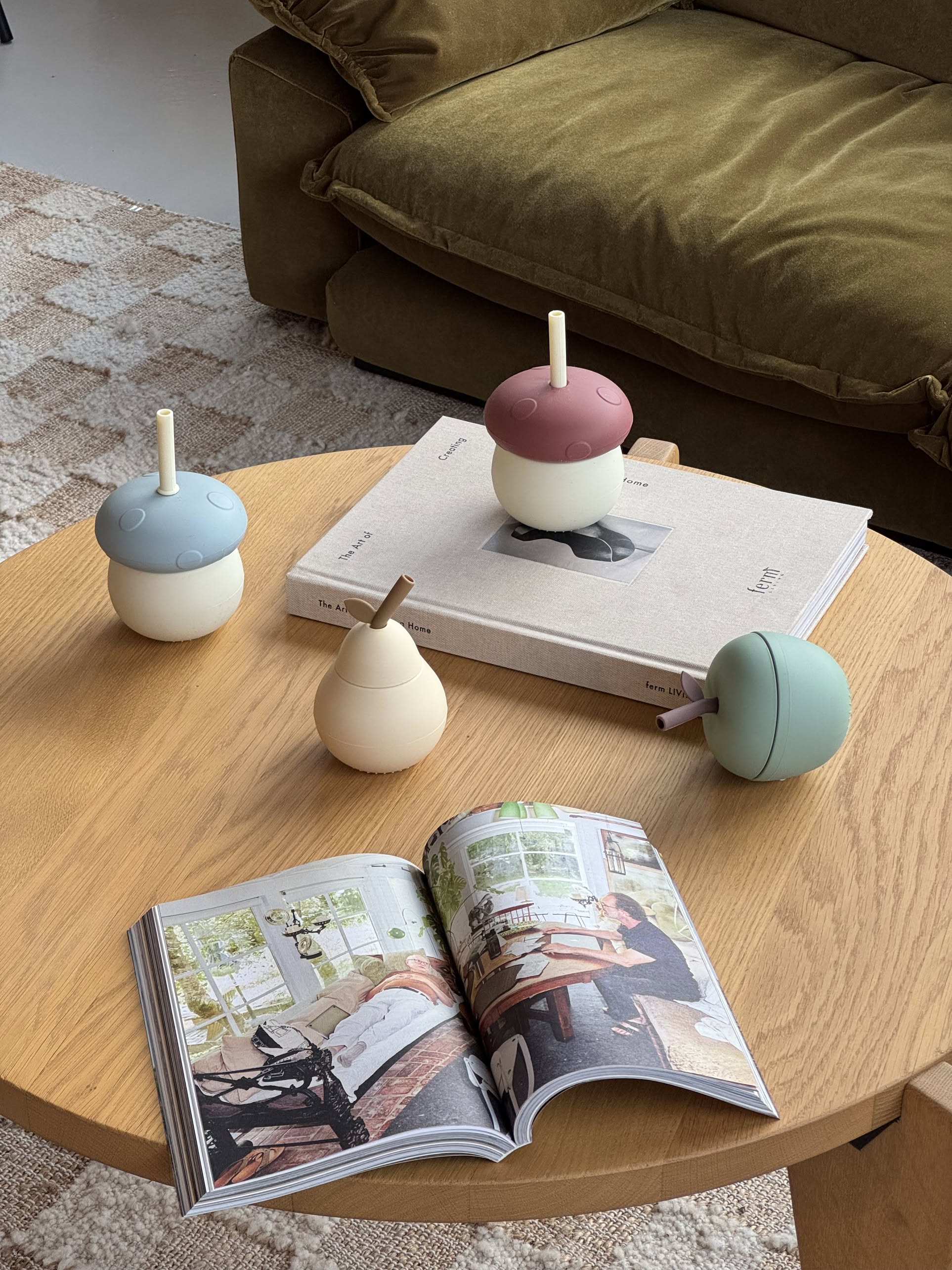 Four OYOY Living Design Pear Cups in Vanilla, each with a silicone acorn shape and straw, sit on a wooden coffee table with a closed book and an open interior design magazine. A sofa and rug appear in the background.