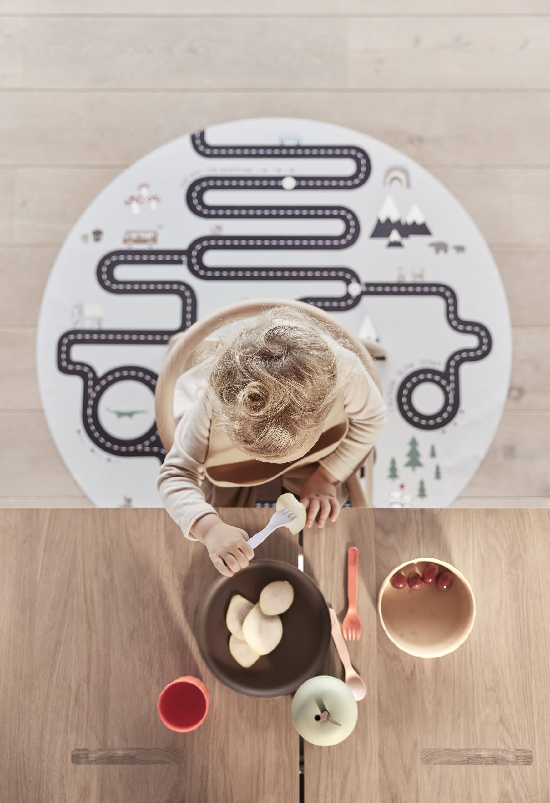 A young child eats sliced fruit from a bowl using the OYOY Living Design Pullo Cutlery Set at a wooden table. On the floor behind them is a round play mat with a road design, and other eco-friendly PLA bowls and utensils are also on the table.