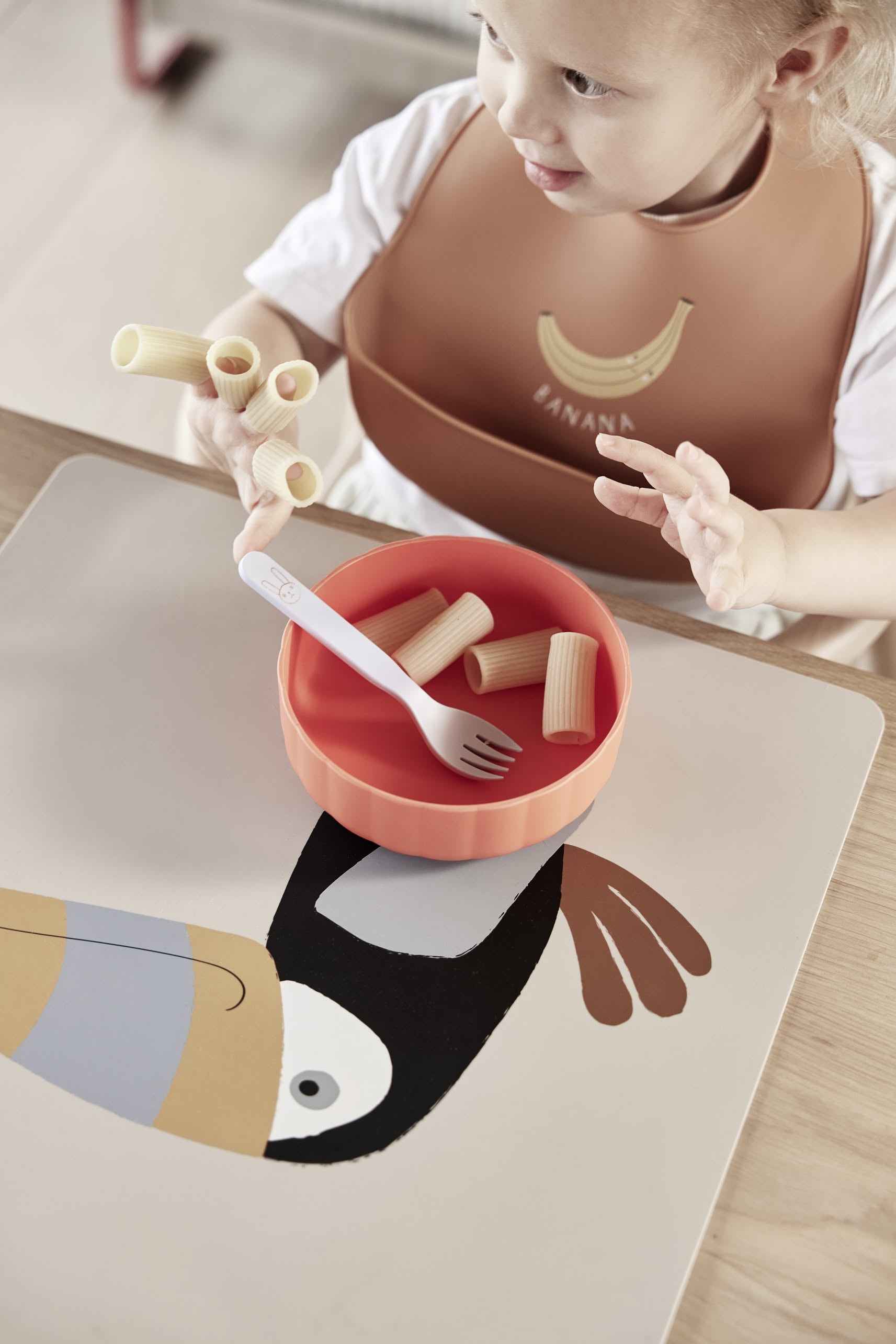 A young child sits at a table holding rigatoni, with a red OYOY Living Design Pullo Cutlery Set and bowl made from eco-friendly PLA on a toucan-illustrated placemat.