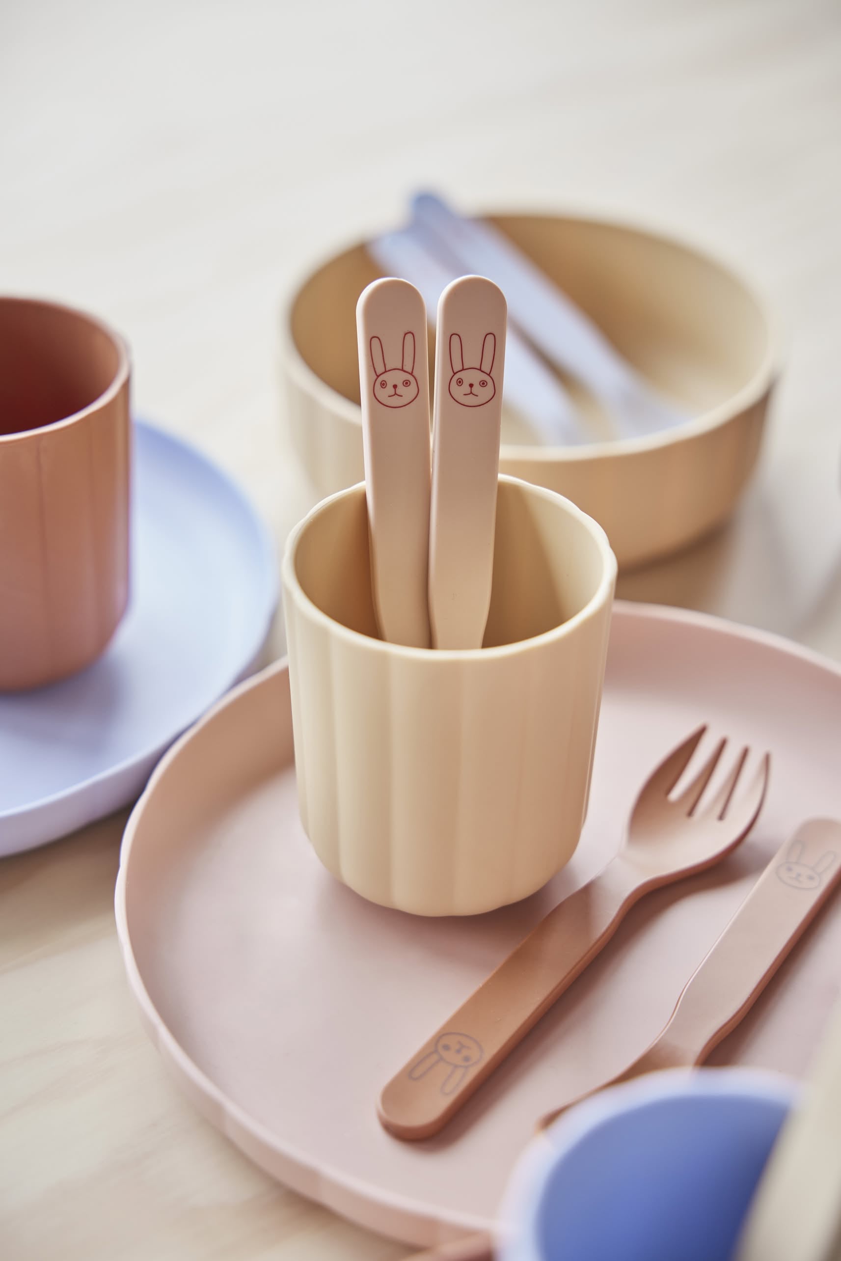 The OYOY Living Design Pullo Cutlery Set features two beige toddler utensils with bunny faces, surrounded by matching pastel plates and bowls made from eco-friendly PLA, beautifully arranged on a light wooden surface.