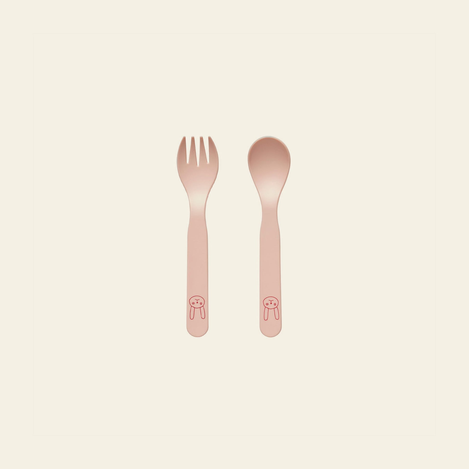 The OYOY Living Design Pullo Cutlery Set by OYOY Living Design is a pink toddler fork and spoon set made from eco-friendly PLA, with simple animal face drawings on the handles, placed side by side on a light background.