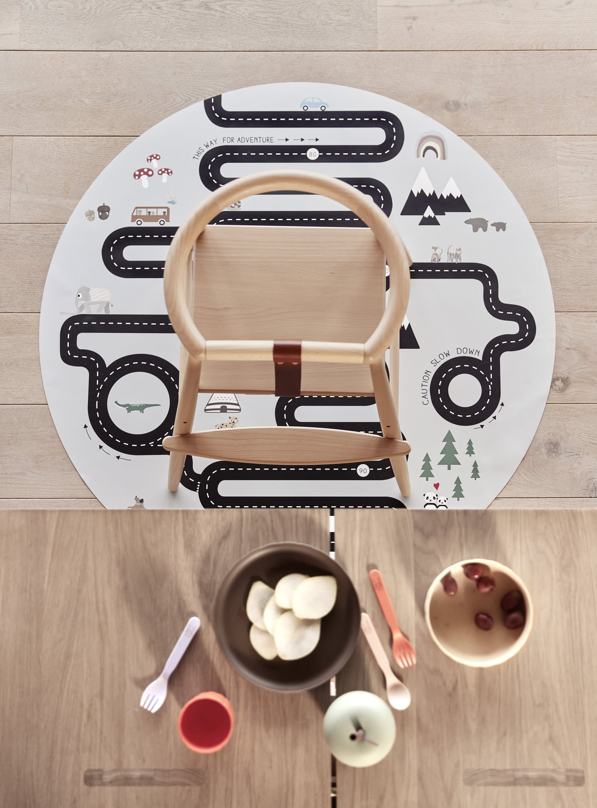 A wooden childs chair sits on a round play mat with roads and nature. In front, an OYOY Living Design Pullo Cutlery Set, a bowl of chips, cherries, forks, cup, and eco-friendly PLA plate rest on a wooden table.