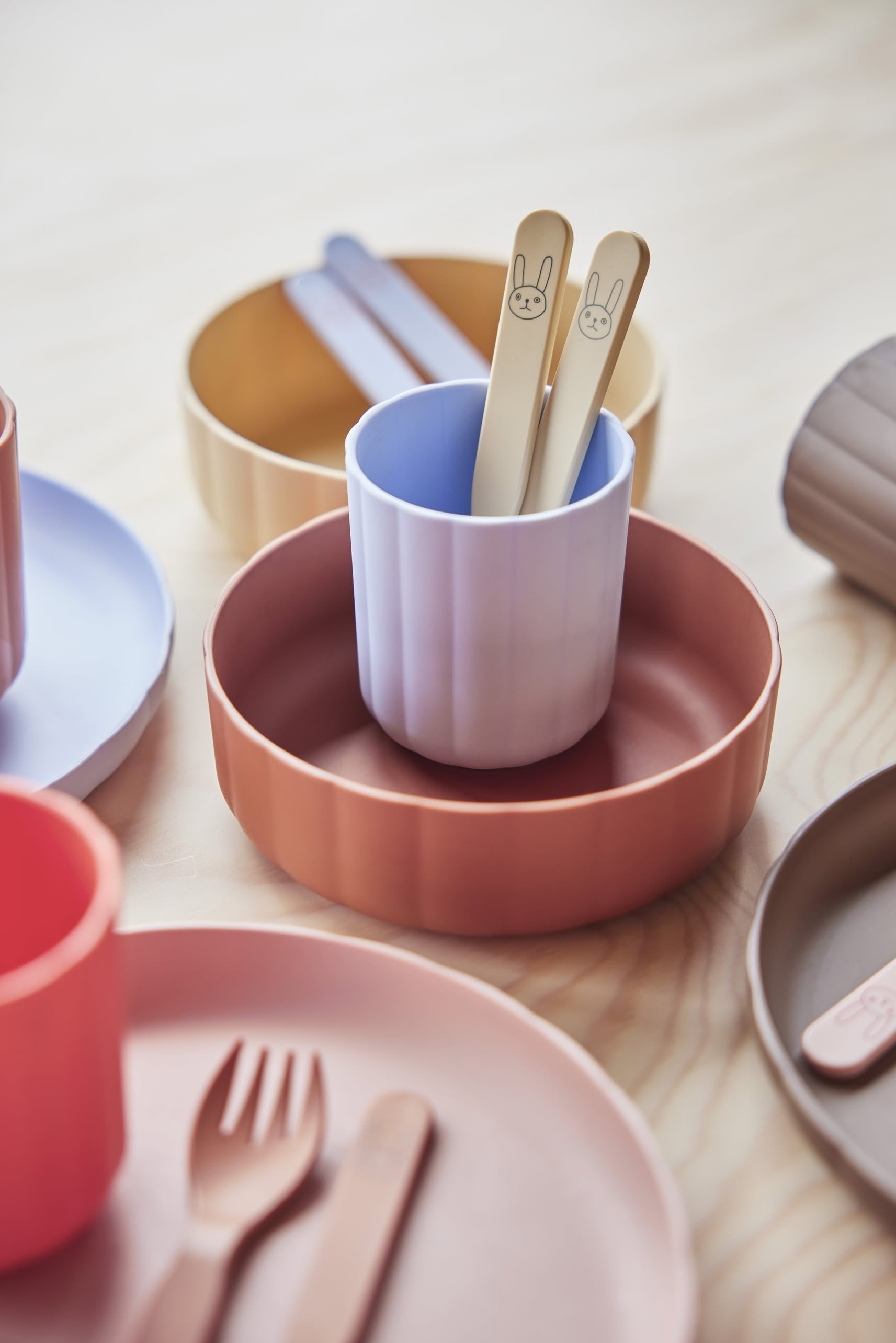 OYOY Living Design Pullo Cutlery Set spoons with bunny faces sit in a purple cup inside a terracotta bowl, surrounded by colorful OYOY Living Design plates, cups, and utensils arranged on a light wooden surface with pastel tableware in the background.
