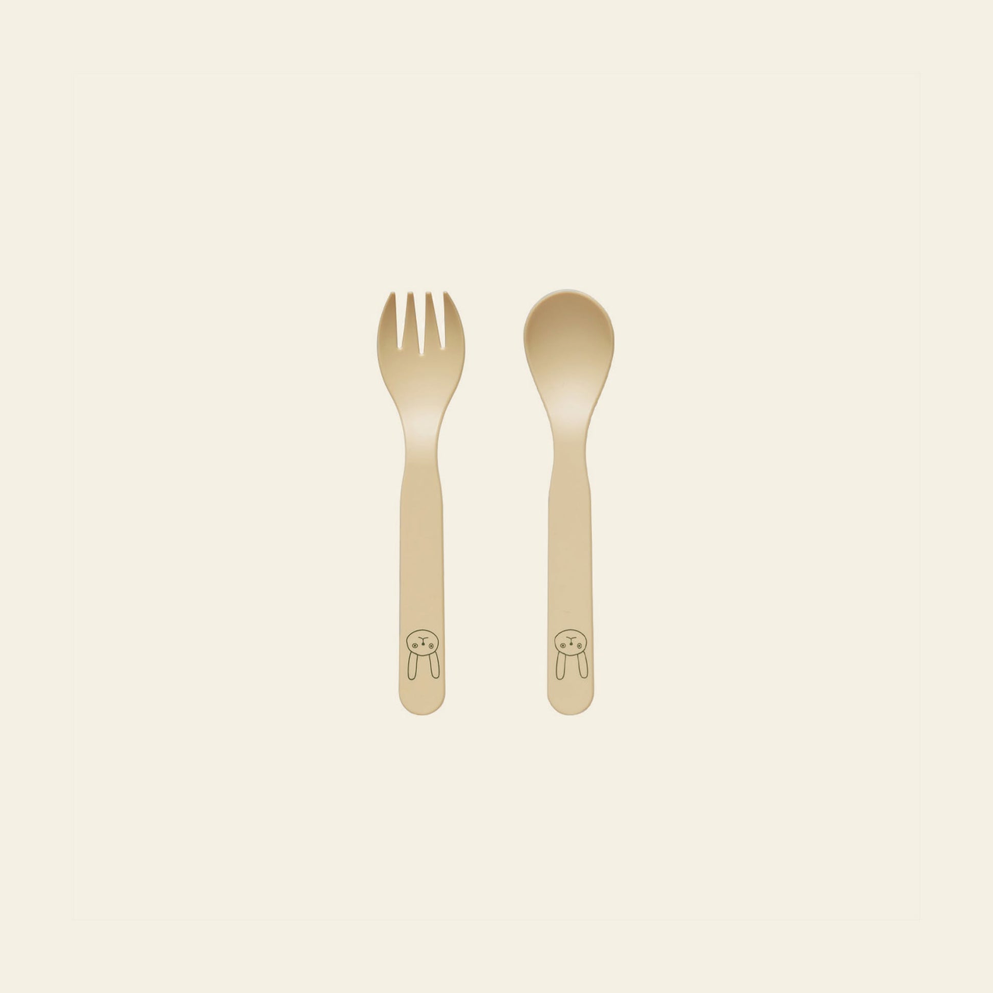 The OYOY Living Design Pullo Cutlery Set for toddlers features a beige fork and spoon made from eco-friendly PLA, rounded handles, and a cute bunny face near the base, displayed on an off-white background.