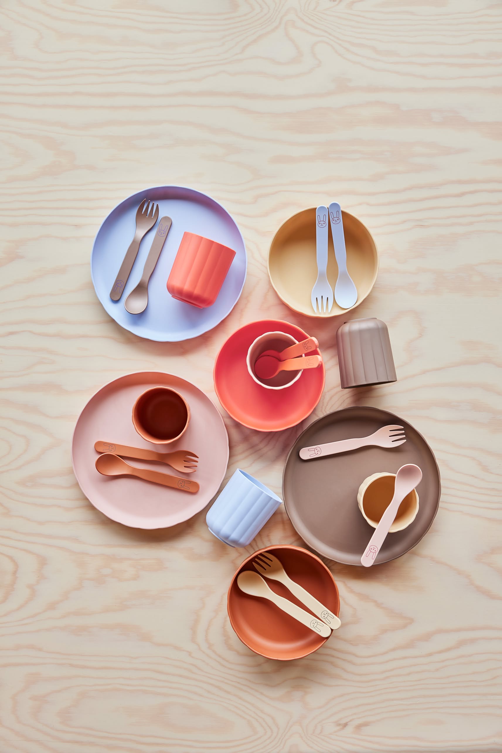 OYOY Living Design Pullo Cutlery Set features colorful, eco-friendly PLA plates, bowls, cups, and toddler cutlery in pastel orange, pink, purple, yellow, and brown arranged neatly on a light wood-grain surface.