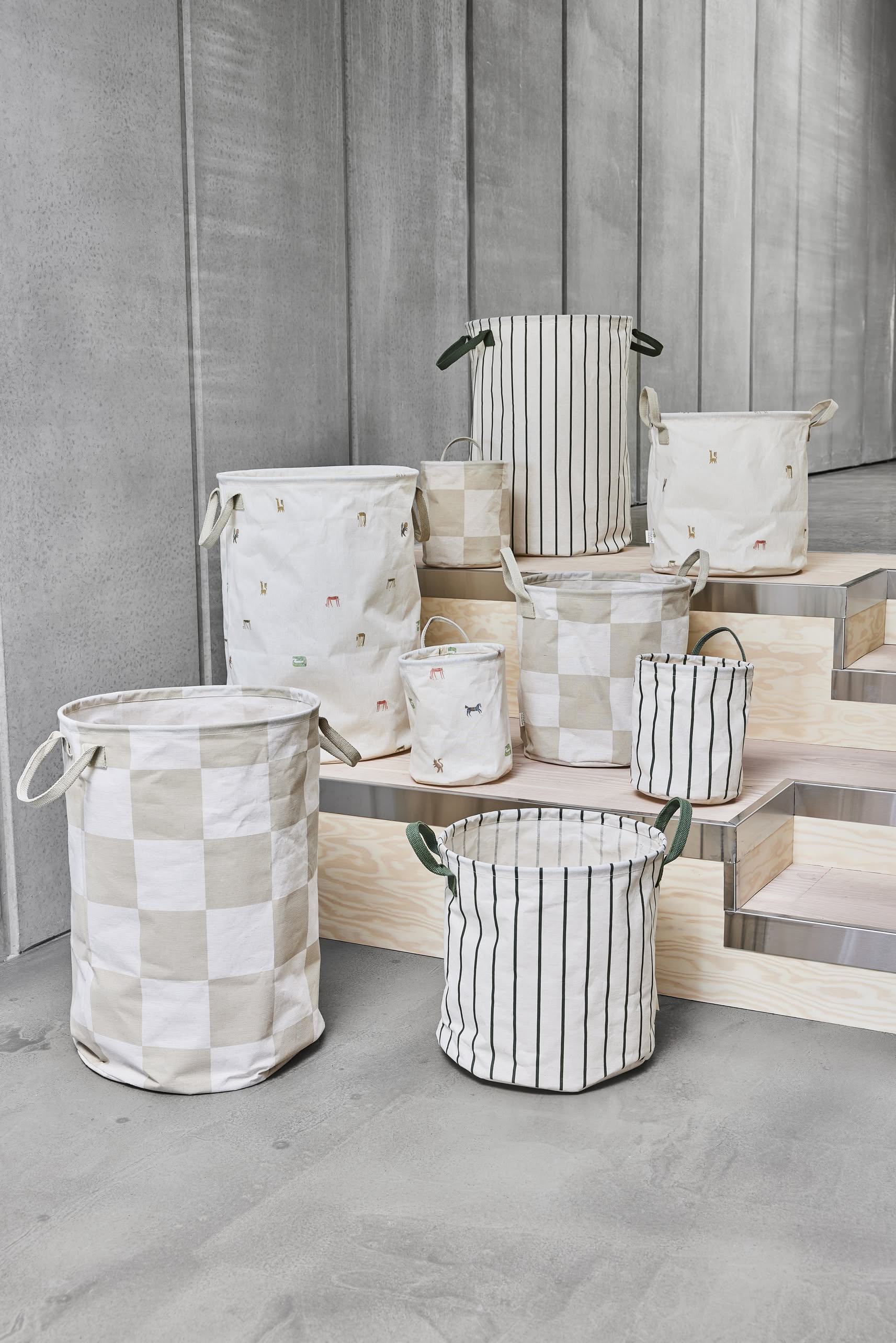 A set of eight OYOY Living Design Raita Laundry & Storage Baskets in medium size, featuring assorted stripes, checks, and animal motifs, is displayed on light wooden steps against a concrete wall.