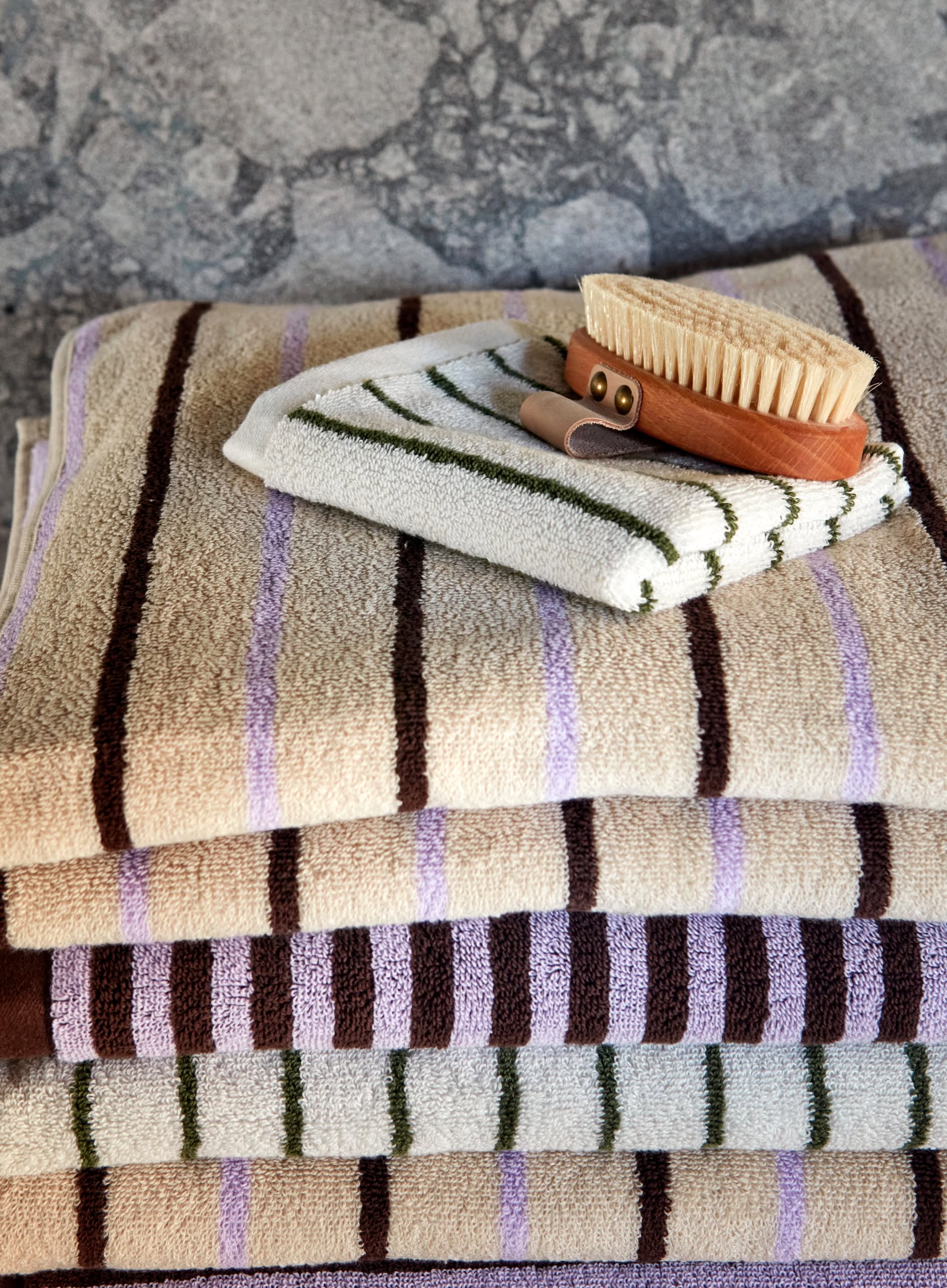 A stack of striped OYOY Living Design Raita Wash Cloths in beige, brown, and lavender sits on a surface, topped with an organic cotton Terry wash cloth and a wooden bath brush with natural bristles.