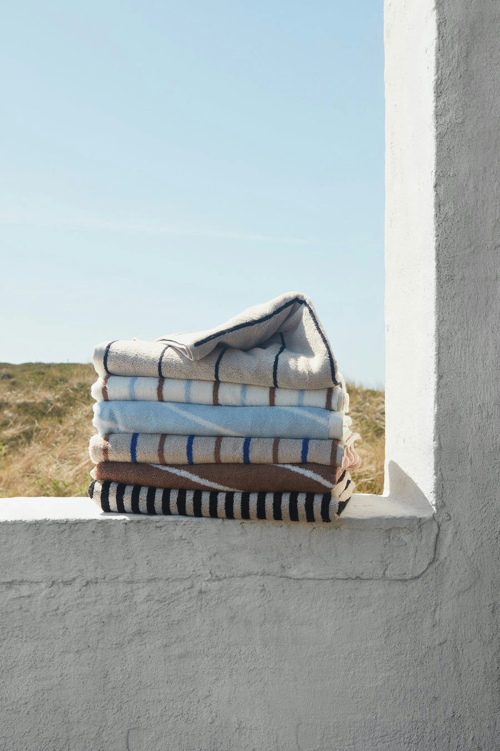 A pack of two OYOY Living Design Raita Wash Cloths, made from organic cotton terry with checkered and striped designs, rests on a white windowsill overlooking a grassy field and blue sky.
