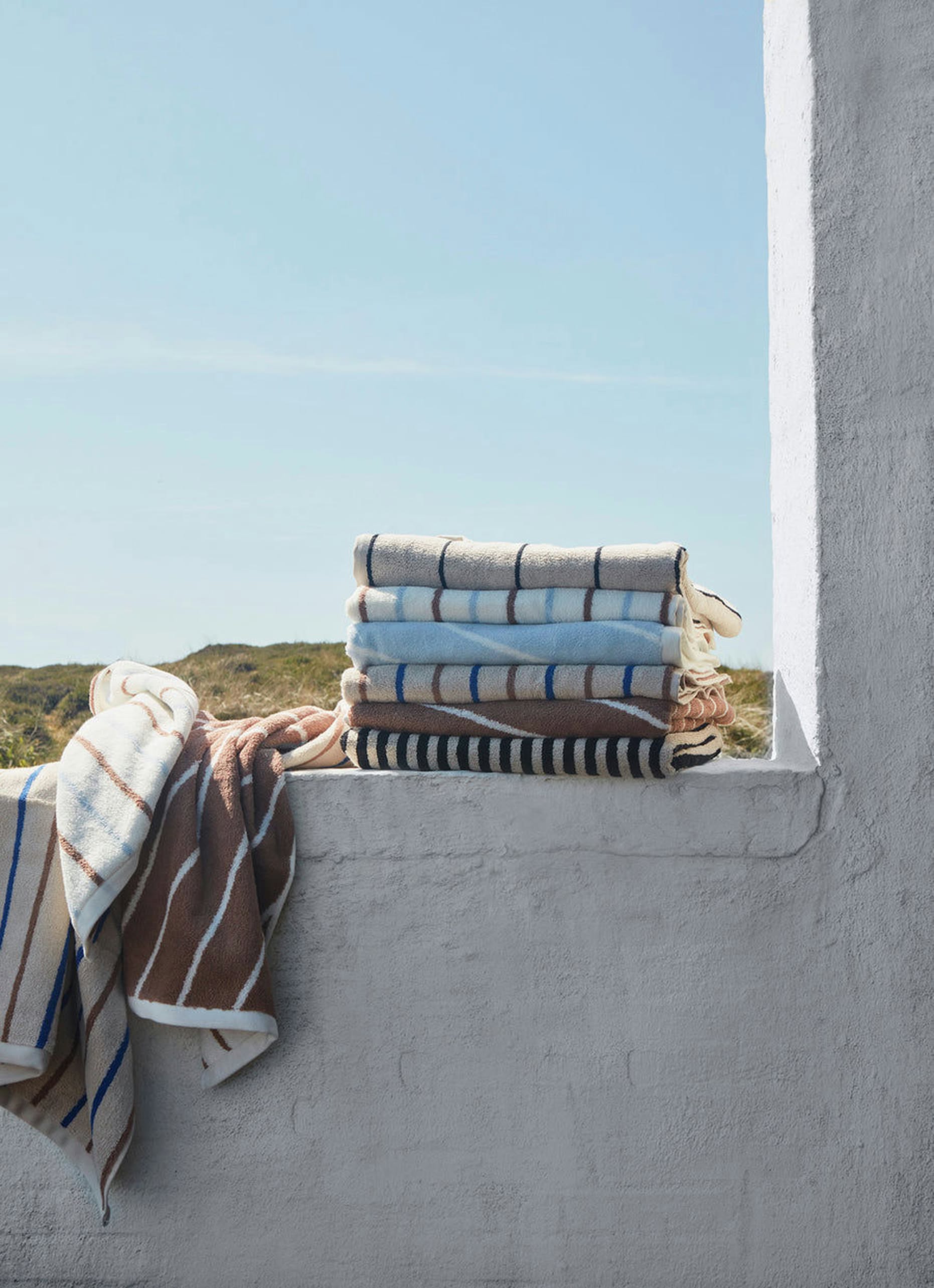 A pair of OYOY Living Design Raita Wash Cloths, crafted from organic cotton terry in blue and beige stripes, rests on a white ledge outdoors with two cloths draped nearby against a lush green landscape.