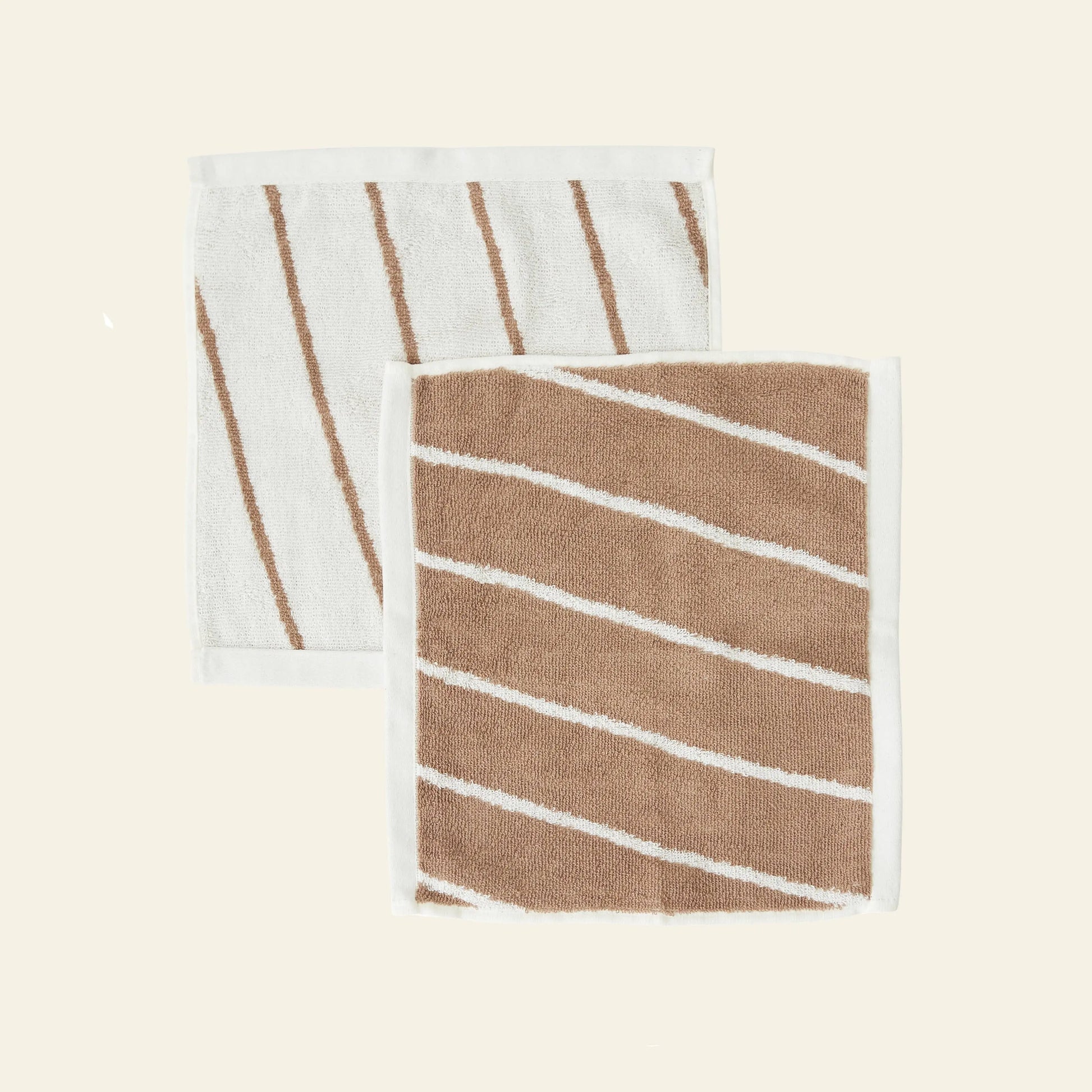 The OYOY Living Design Raita Wash Cloth, Pack of 2, features organic cotton terry cloths with diagonal stripes—one mainly white with slim brown stripes, one mainly brown with slim white stripes—each bordered in white on a light beige background.