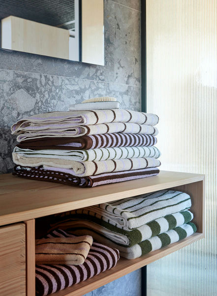A stack of OYOY Living Design Raita Wash Cloths, Pack of 2, in brown, green, and white stripes sits on a wooden shelf in a modern bathroom with a stone wall and frosted glass panel.