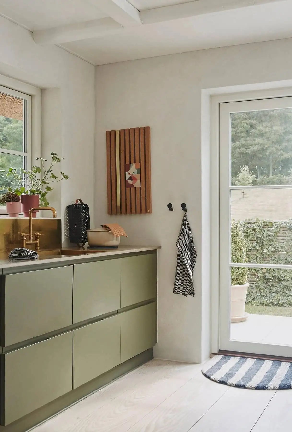 A modern kitchen with olive green cabinets, brass sink, potted plants, striped rug, and OYOY Living Design Stringa Mini Towels hanging on wall hooks. A large glass door brings in light while wooden art decorates the white wall.