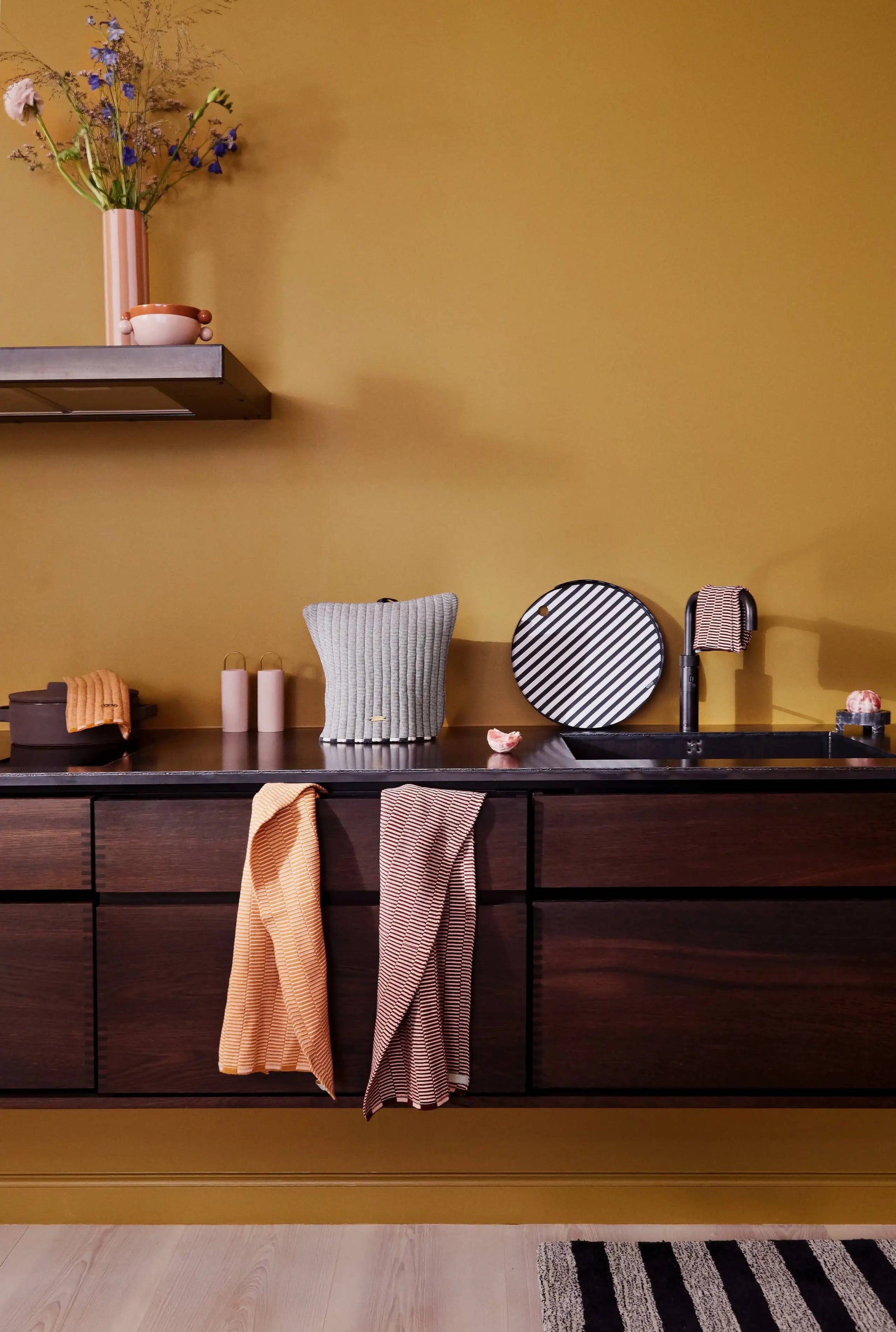 A modern kitchen with dark wood cabinets, black countertop, and matte black sink features OYOY Living Design Stringa Mini Towels in knitted organic cotton hanging from the cabinet, set against a mustard yellow wall with vases and dishes.