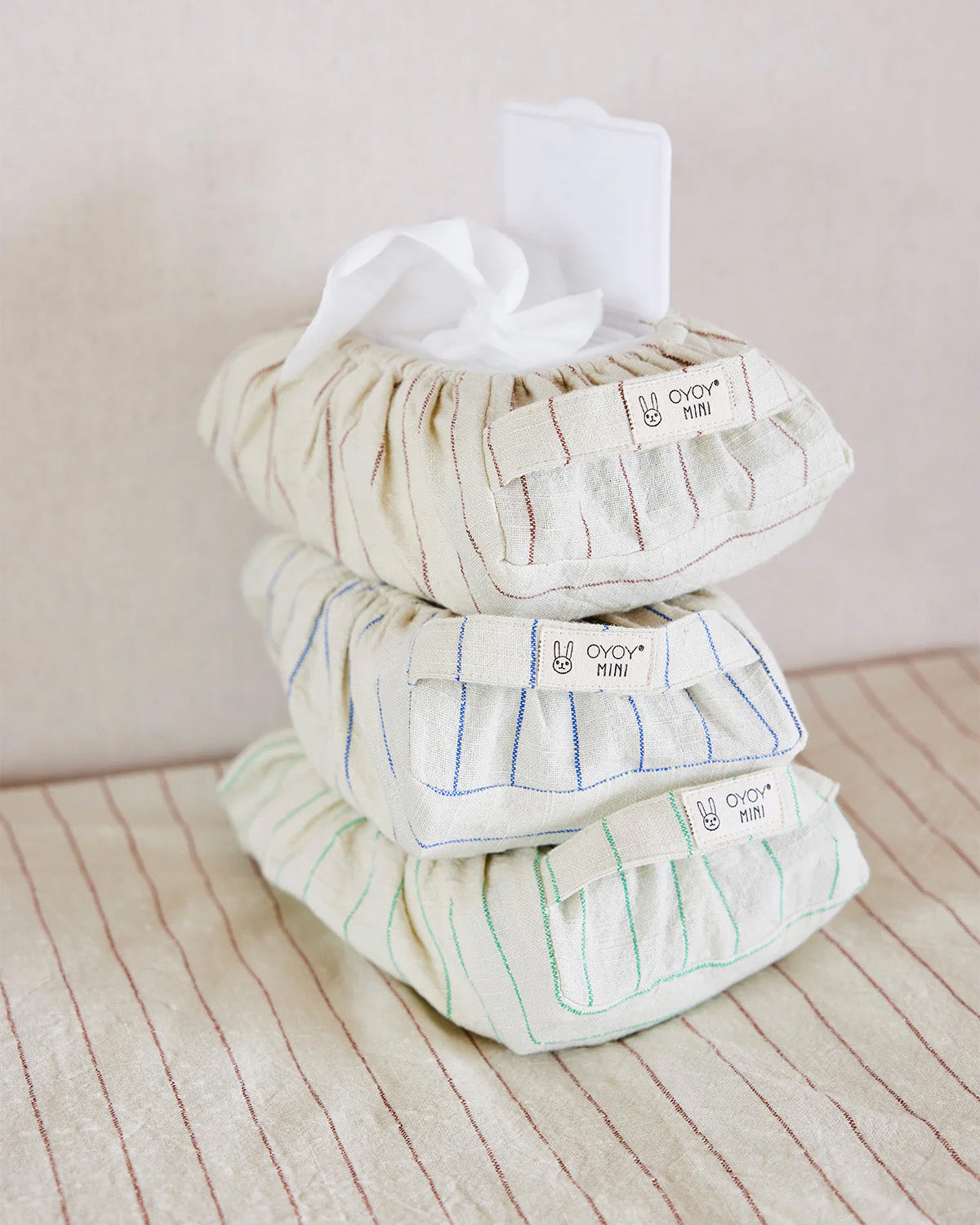 Three striped fabric pouches labeled OYOY Living Design are stacked, with the top pouch—the Nutmeg Wet Wipes Cover—open to reveal white wipes. The background shows matching striped fabric.
