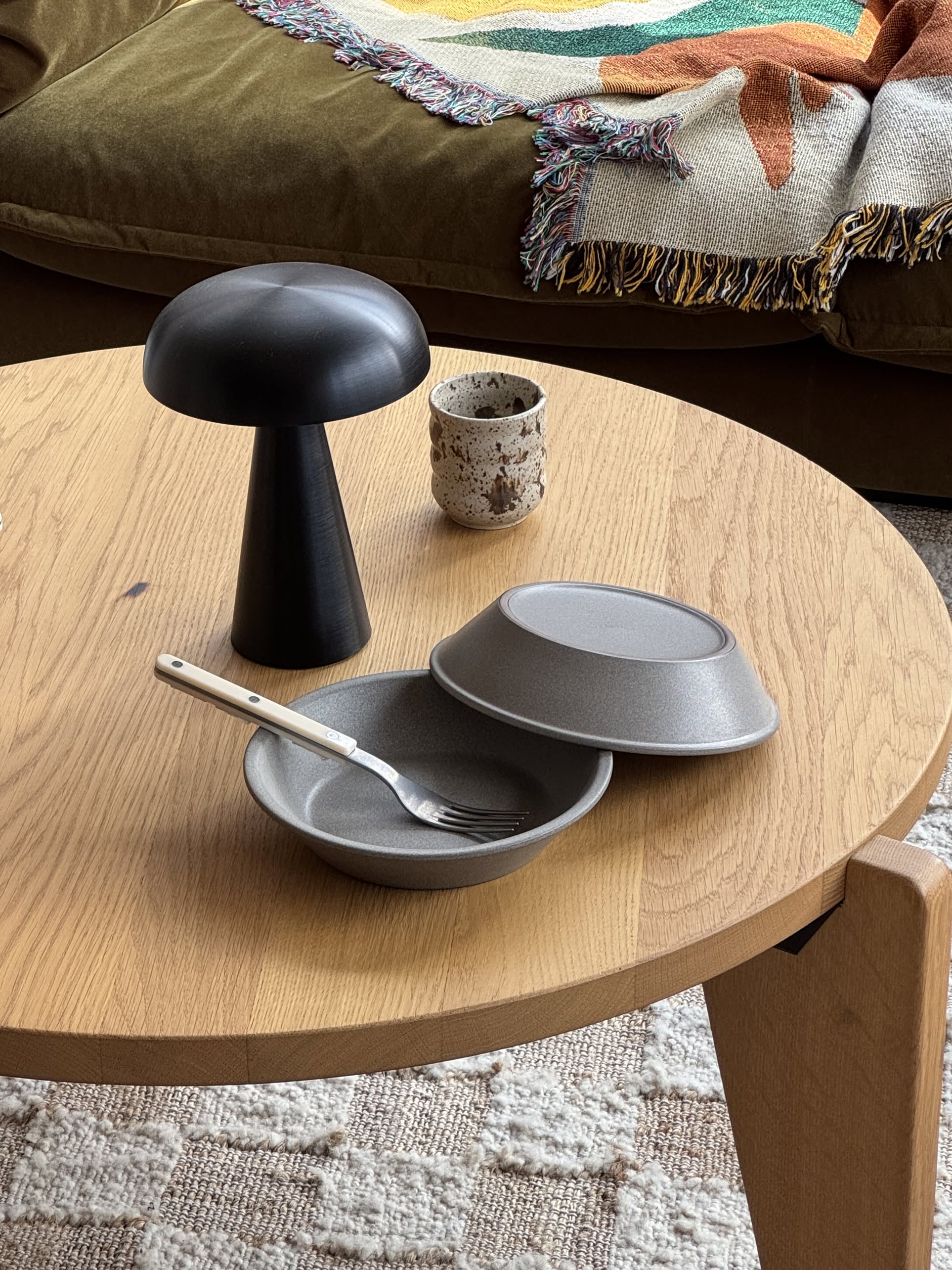A wooden coffee table features OYOY Living Design Yuka Deep Plate, Set of 2 - Stone; one bowl is upside down. A fork, a small black mushroom lamp, and a speckled cup sit nearby. A patterned blanket covers the green couch in the background.