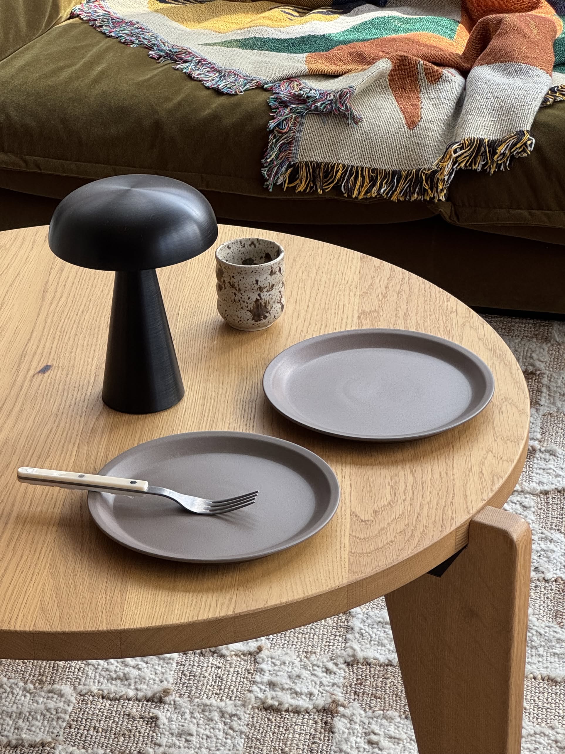 A round wooden table with a modern setting: two gray OYOY Living Design Yuka Dinner Plates (Set of 2 - Stone) with fork and knife, a black mushroom lamp, speckled ceramic cup, and a fringed blanket draped over a brown couch in the background.
