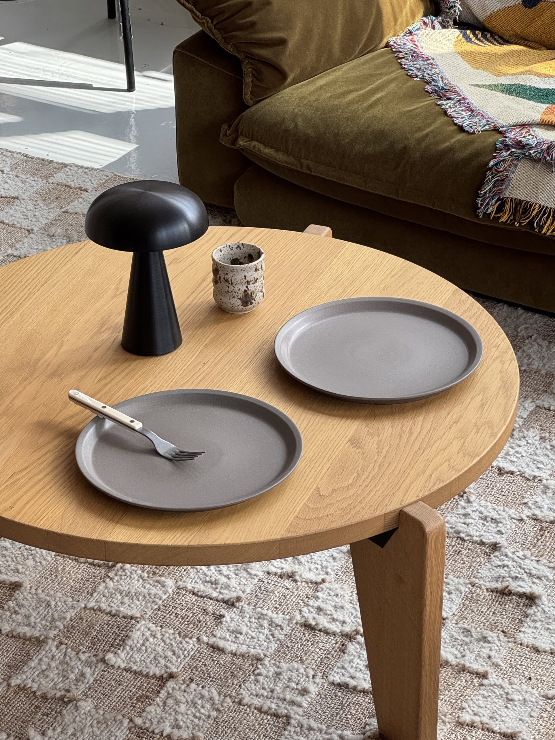 A round wooden coffee table with two OYOY Living Design Yuka Dinner Plates in Stone, a fork on one, a small ceramic cup, and a black mushroom-shaped lamp. A green sofa with a colorful blanket sits behind on a patterned rug.