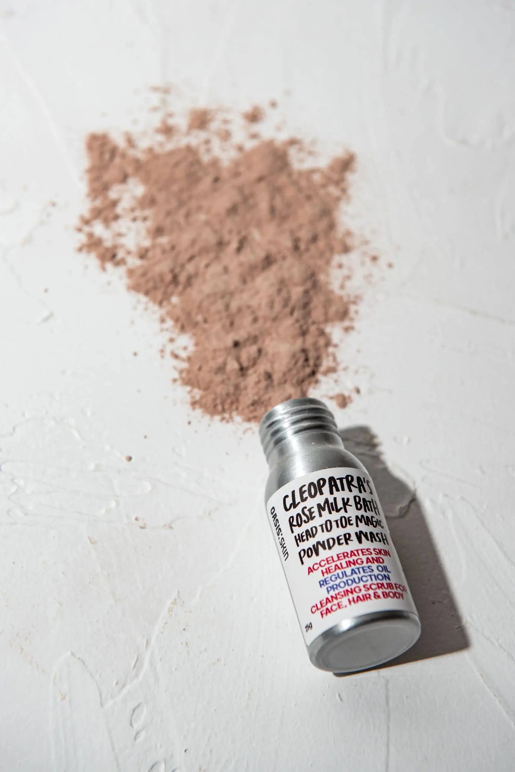 A small open bottle labeled Oasis Beauty Kitchen Head to Toe Magic Powder Wash, 25g lies on a white surface with a pile of pinkish powder, made from plant-based ingredients and organic essential oils, spilled behind it. Suitable for face, hair, and body care.