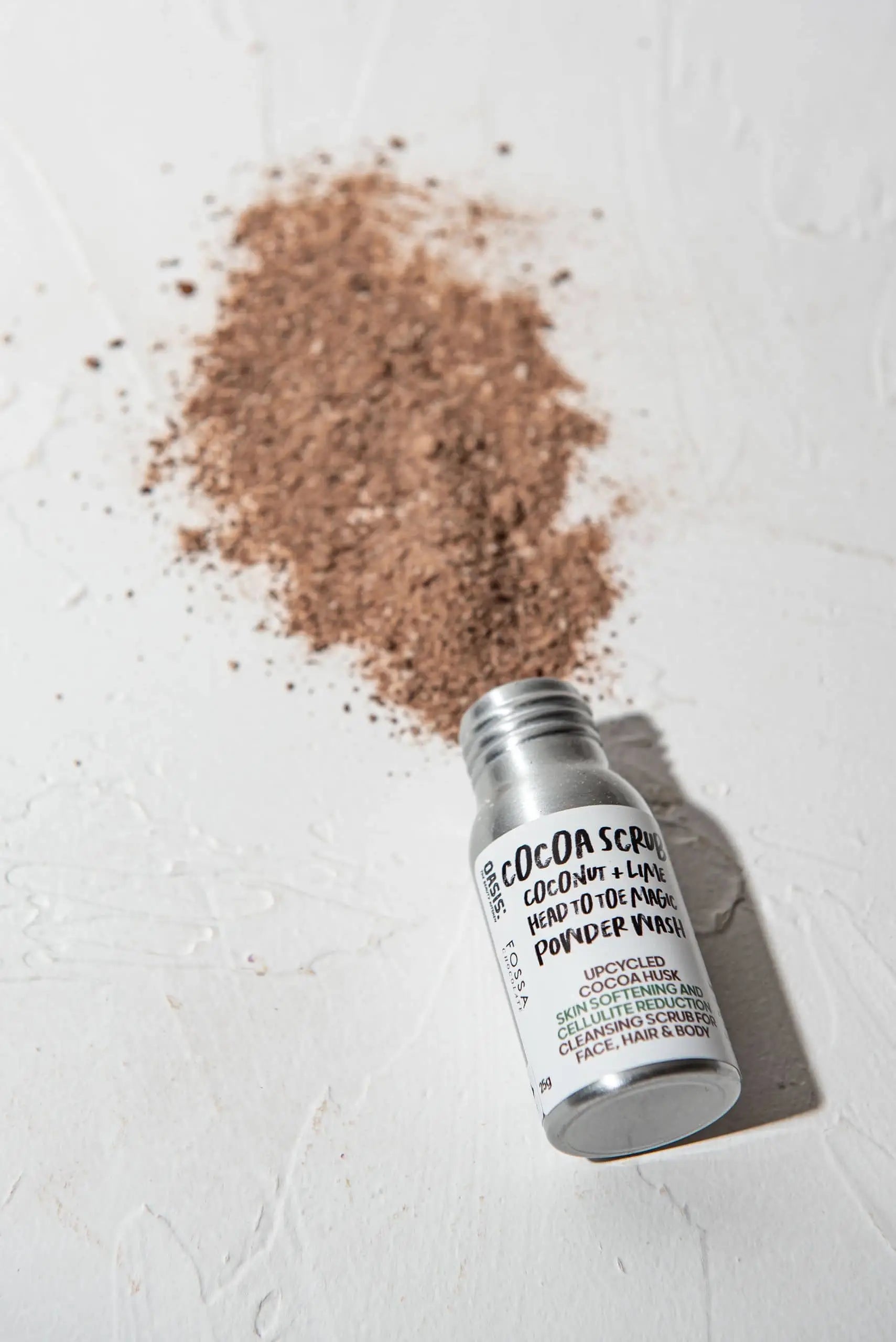 A small silver bottle of Oasis Beauty Kitchen Head to Toe Magic Powder Wash, 25g, lies on its side, spilling fine brown vegan powder onto a textured white surface.