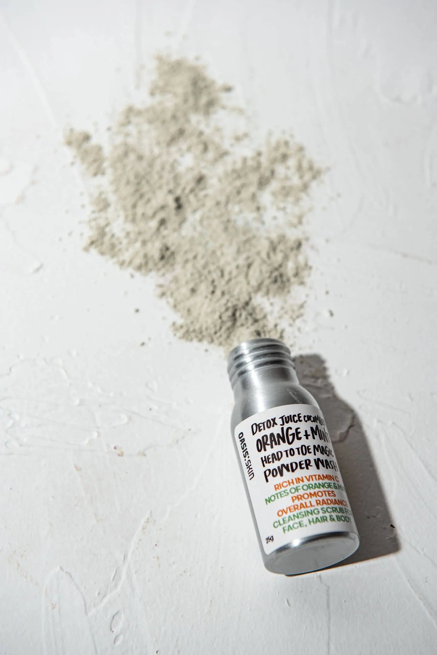 A small bottle labeled Oasis Beauty Kitchen Head to Toe Magic Powder Wash, 25g, rests on a white surface with some light gray powder spilled—crafted by Oasis Beauty Kitchen for a gentle, effective vegan wash using plant-based ingredients.