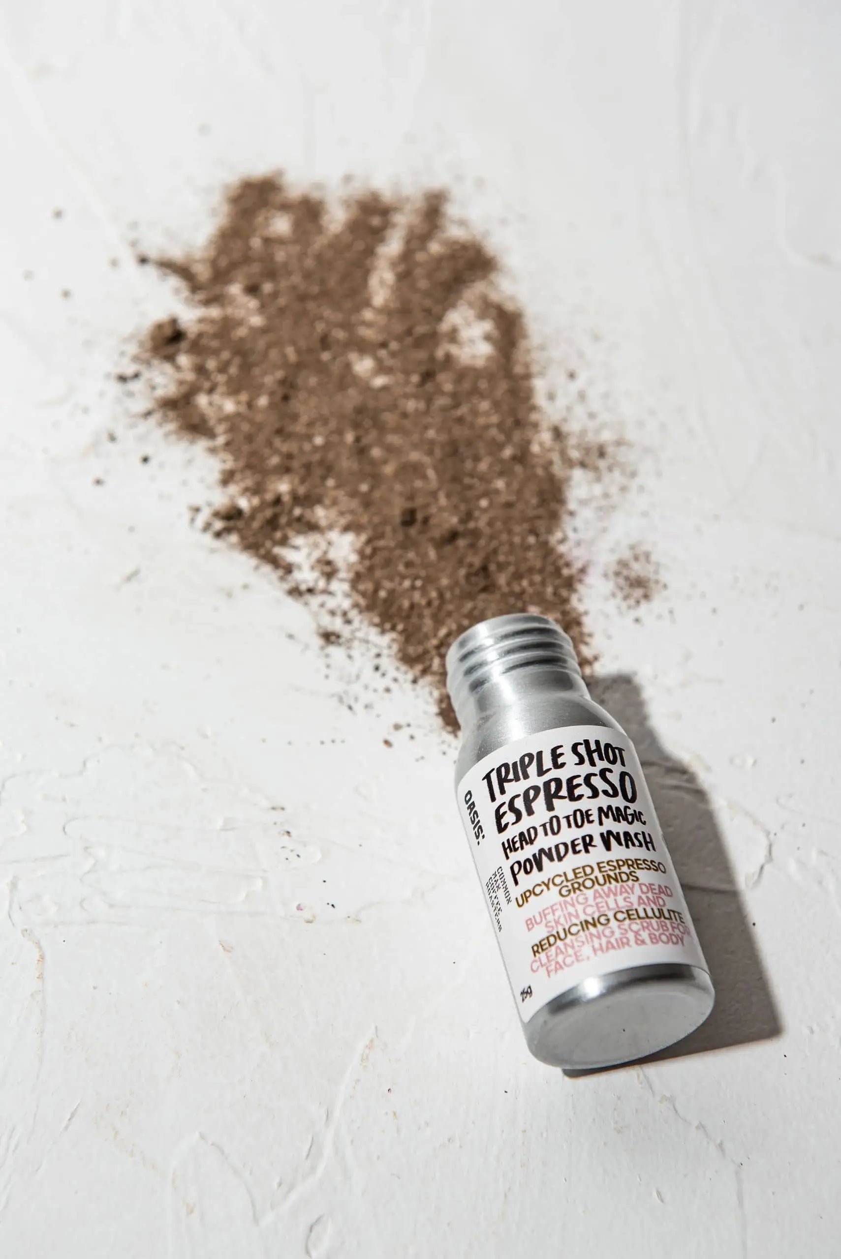 A small container of Oasis Beauty Kitchen Head to Toe Magic Powder Wash, 25g, lies on its side, plant-based vegan powder spilled out onto a white surface.