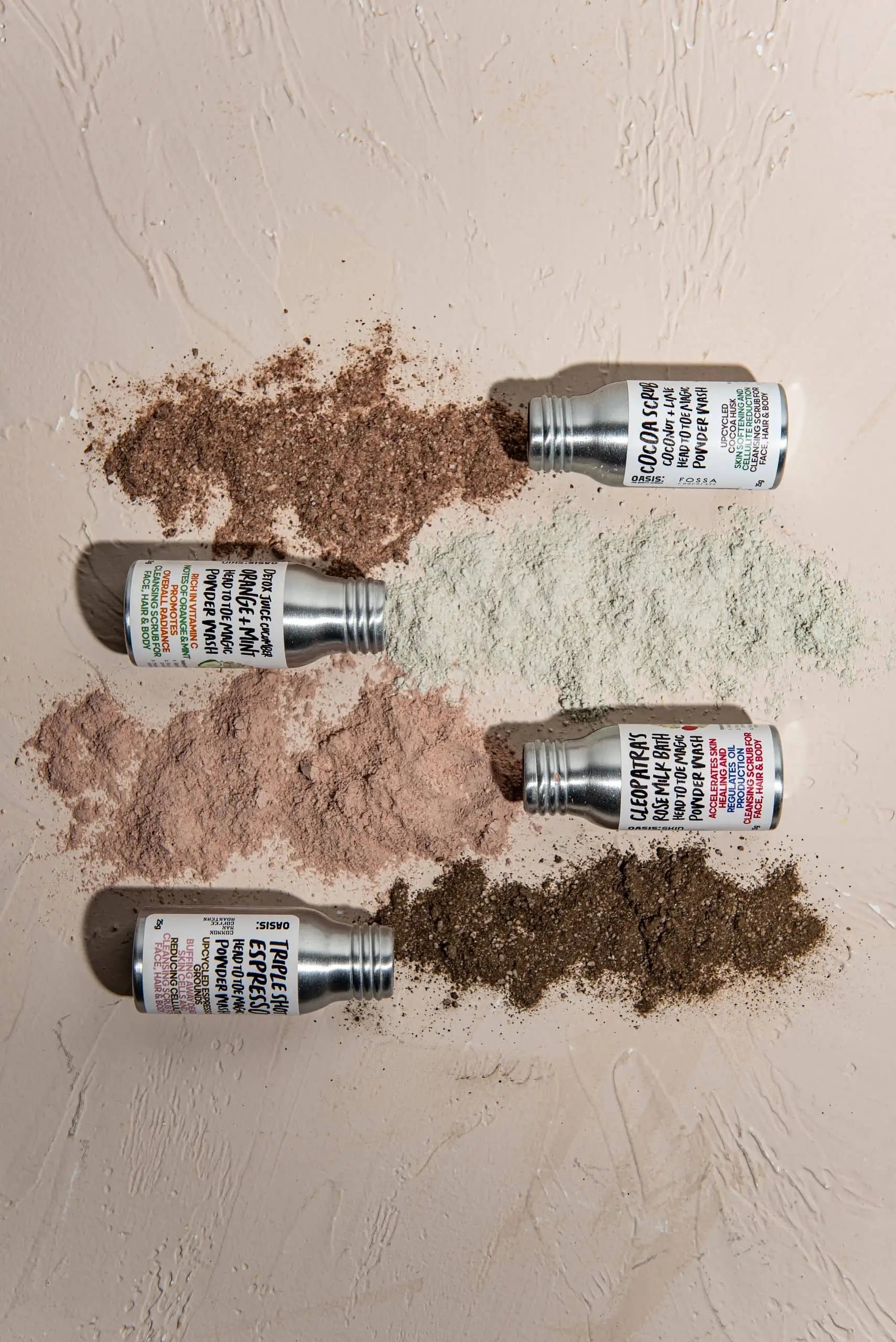 Four Oasis Beauty Kitchen Head to Toe Magic Powder Wash bottles (25g each) lie on a textured surface, pouring brown, green, pink, and dark brown vegan powders. Each features a colorful label showcasing plant-based ingredients.