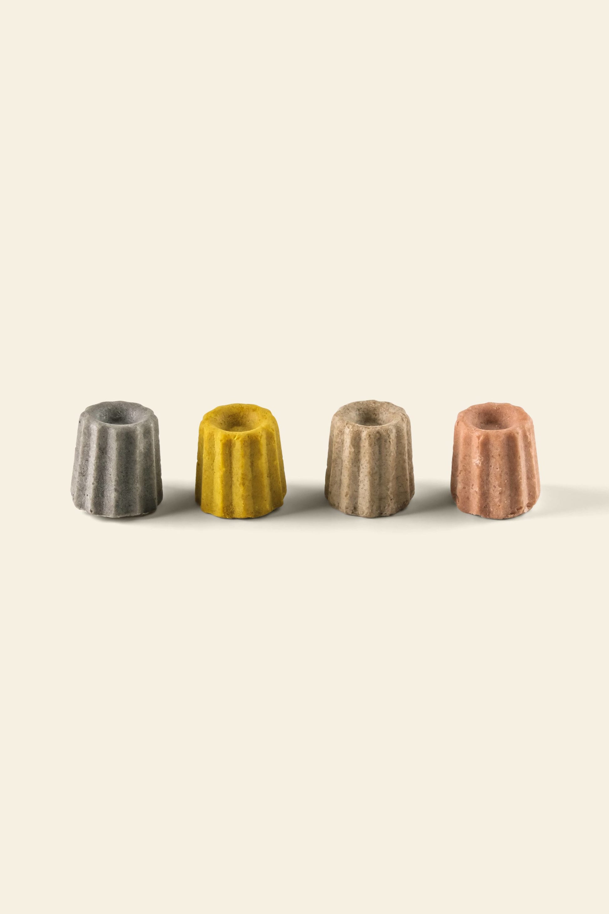 Four small, fluted, cylindrical Oasis Beauty Kitchen Miniature Solid Shampoos in gray, yellow, beige, and pink are aligned in a row on a light beige background, each casting a soft shadow.