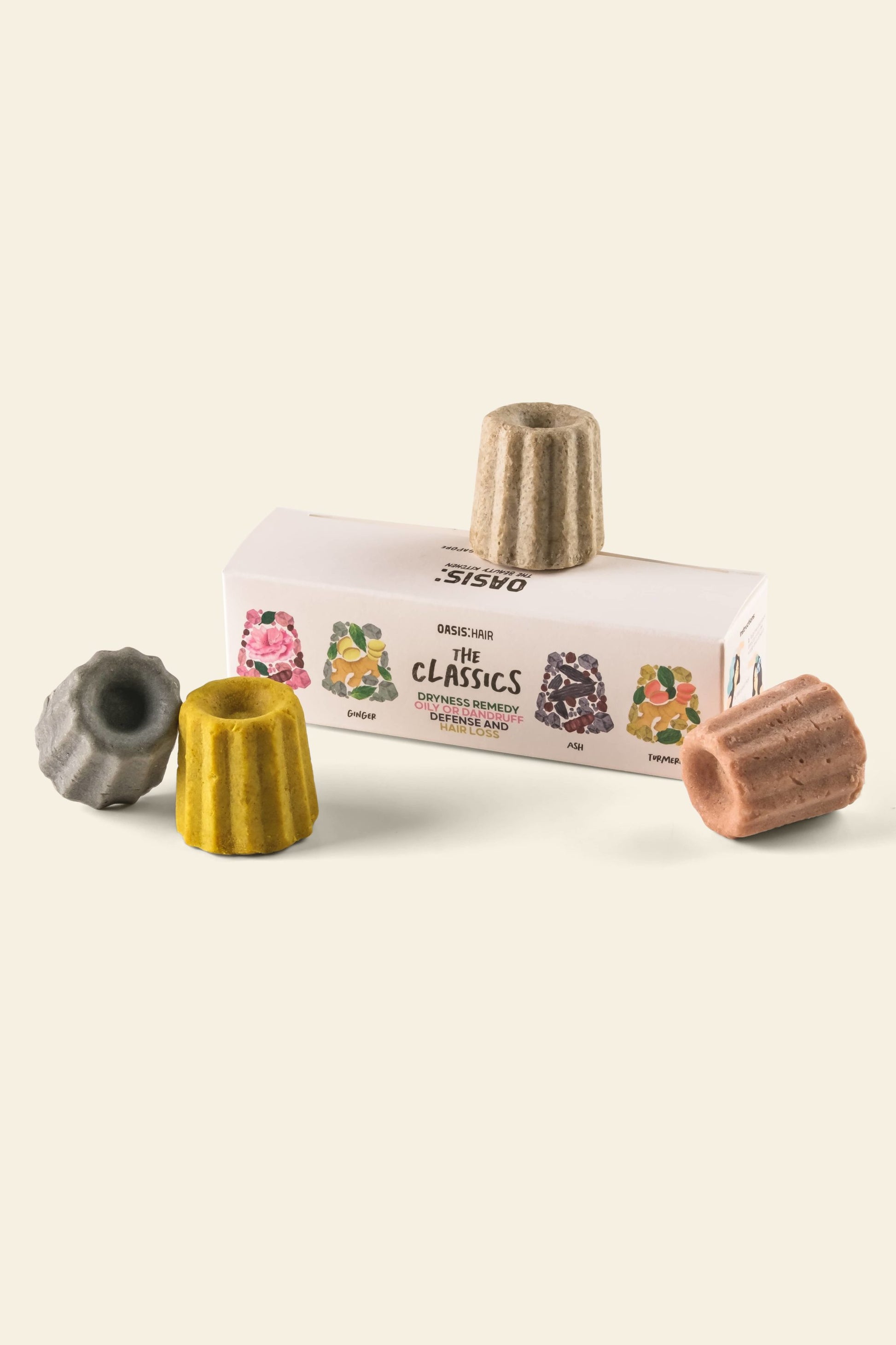 Three colorful, canelé-shaped Oasis Beauty Kitchen Miniature Solid Shampoos sit beside and atop a rectangular box labeled The Classics from Oasis Beauty Kitchen. The box shows illustrations that match the gray, yellow, brown, and tan shampoos.
