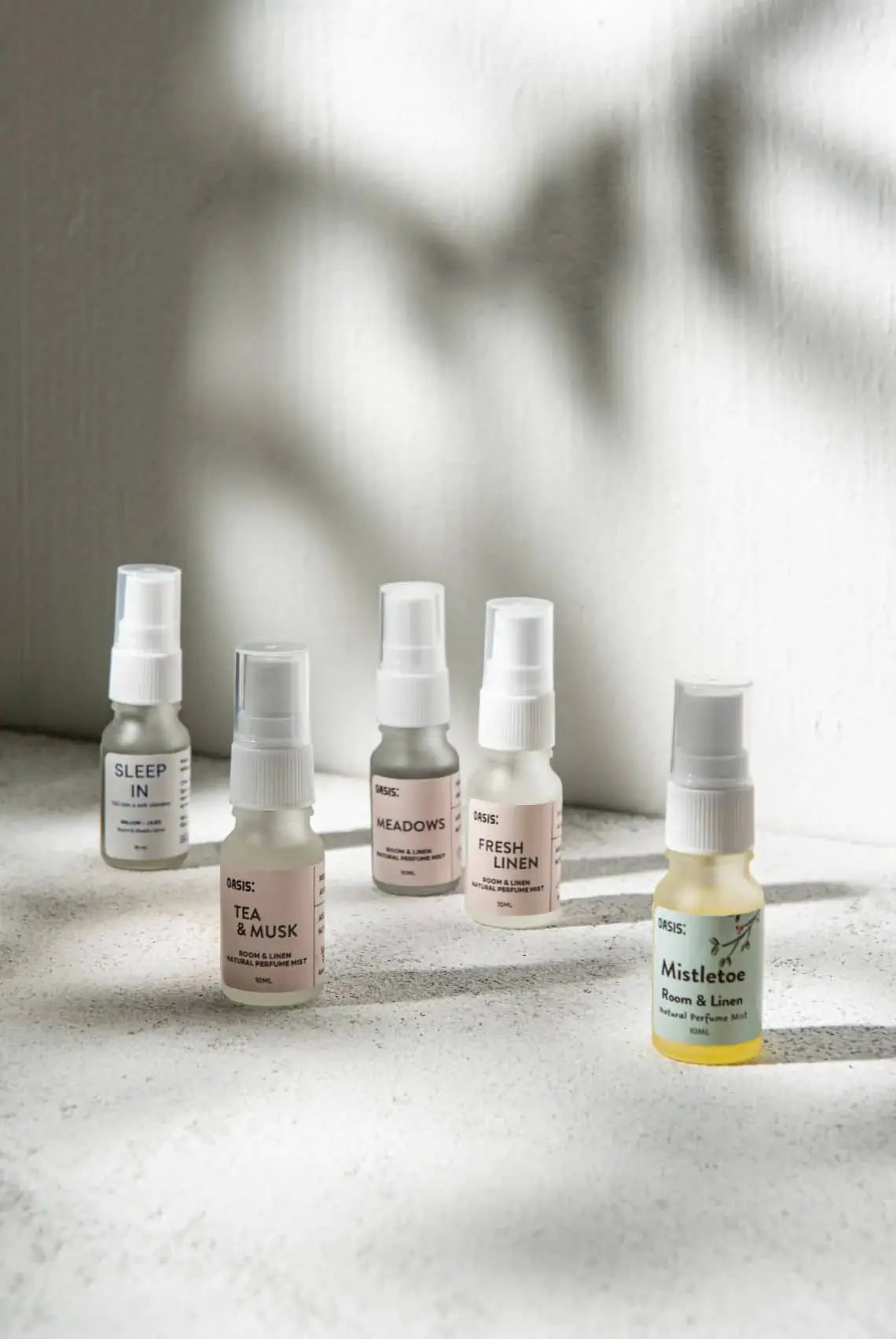 Five small Oasis Beauty Kitchen Room and Linen Mist 10ml bottles sit on a textured white surface in sunlight. Four have neutral labels, while the front right bottle shows a yellow Oasis Beauty Kitchen label. Shadows stretch behind them.