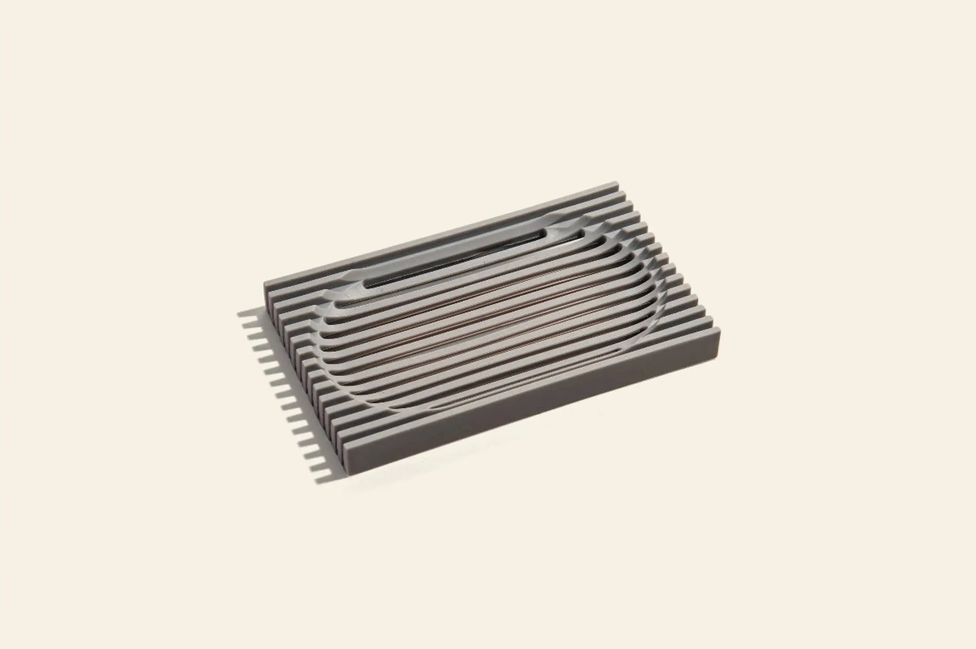 The Oasis Beauty Kitchen The Lattice Soap Dish by Oasis Beauty Kitchen features a modern rectangular design with durable metallic construction and parallel grooves, perfect for organizing soaps on a light beige background.