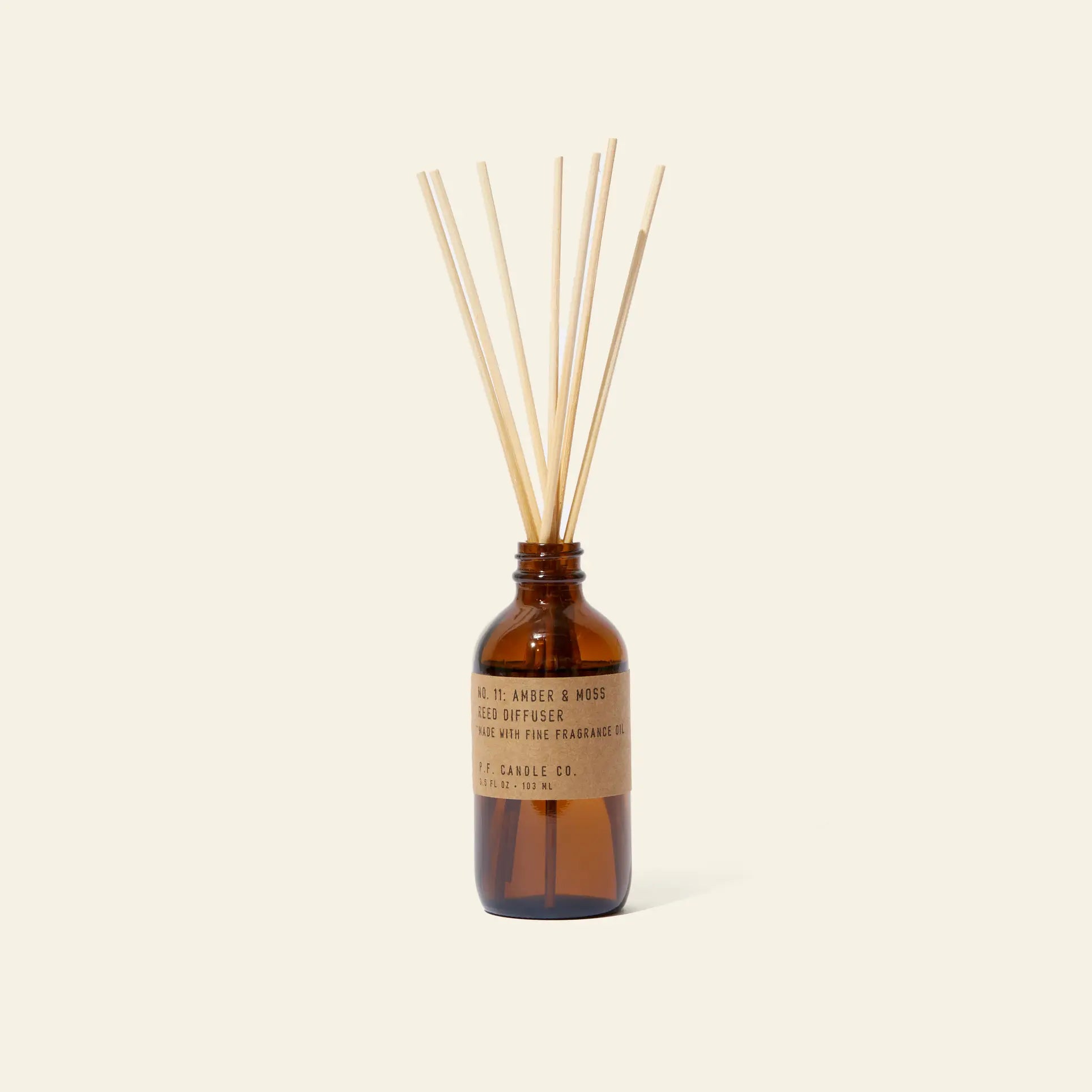 The P.F. Candle Co. 3.5oz Reed Diffuser - Amber & Moss features a brown glass bottle with a kraft label, filled with fragrance oils and several natural-colored diffuser sticks, set against a plain, light background.
