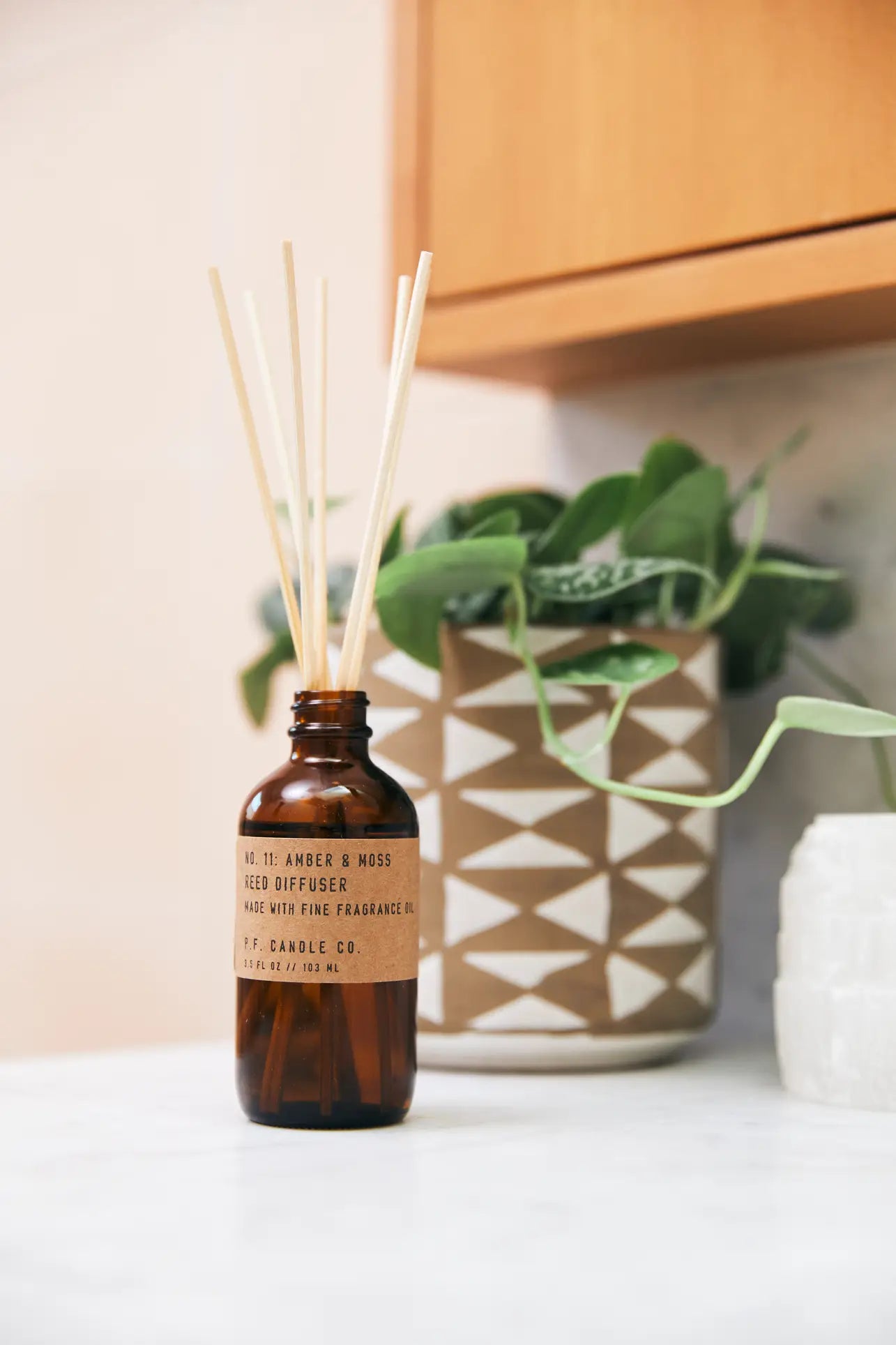 The P.F. Candle Co. 3.5oz Reed Diffuser in Amber & Moss, with wooden reeds and brown glass bottle, sits on a white countertop next to a potted plant and textured container, beneath warm wooden cabinets.